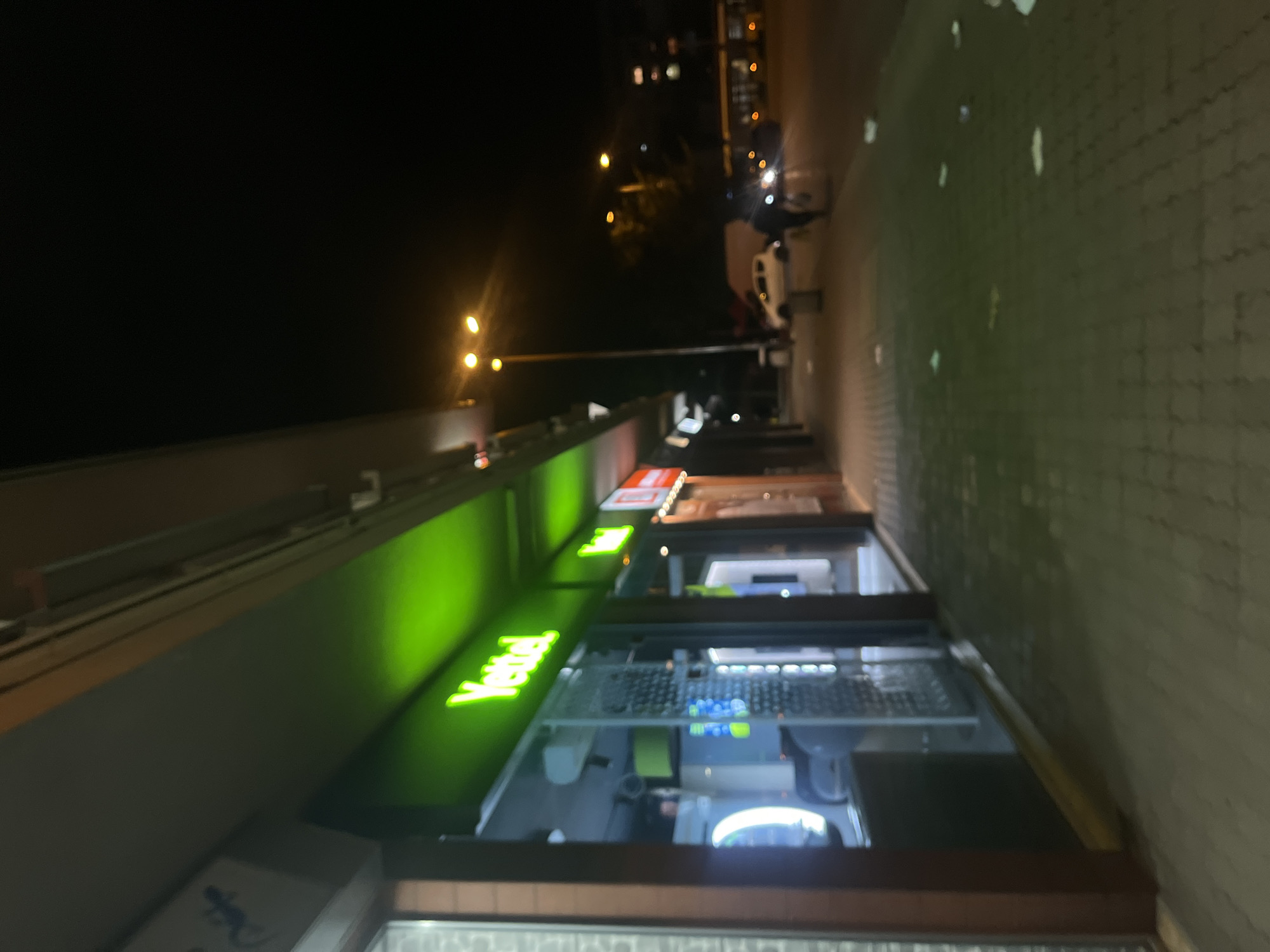 The image depicts a lit "Yettel" store at night in Sofia, Bulgaria. The store front is brightly illuminated with the green "Yettel" logo. Inside the store, computer setups are visible. The street is paved. In the background, there are streetlights and blurred lights of vehicles. The image is taken from a street-level perspective.