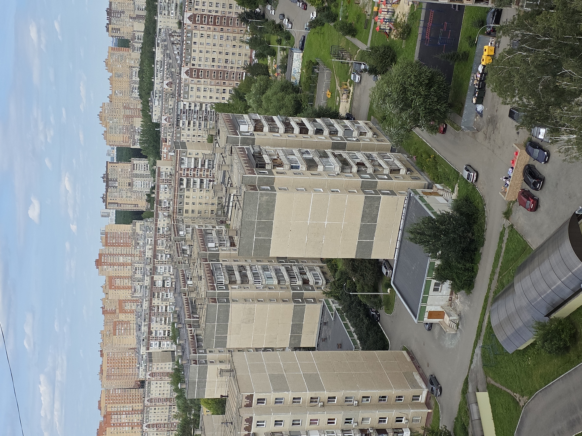 The image depicts a view of a residential area in Chelyabinsk, Russia, characterized by multi-story apartment buildings. The architecture suggests a Soviet-era or post-Soviet urban design, with numerous identical or similar apartment blocks constructed in concrete and brick, painted in muted colors like beige, grey, and some with orange accents on the upper floors. The buildings are densely packed, with balconies visible on many units.
Green spaces, including trees and grass, are interspersed between the buildings, indicating a typical urban layout with attempts to incorporate nature. Several cars are parked on the paved areas and roads surrounding the buildings. A playground with red and blue structures is visible in the mid-ground. In the foreground, a large, curved, dark-colored structure, possibly a covered walkway or bus stop, is partially visible. A cluster of yellow and black trash bins is also present, with some debris around them. The sky is partly cloudy, suggesting daytime. No specific landmarks are identifiable to pinpoint the exact location within Chelyabinsk, but the general appearance is consistent with many residential districts in Russian cities.