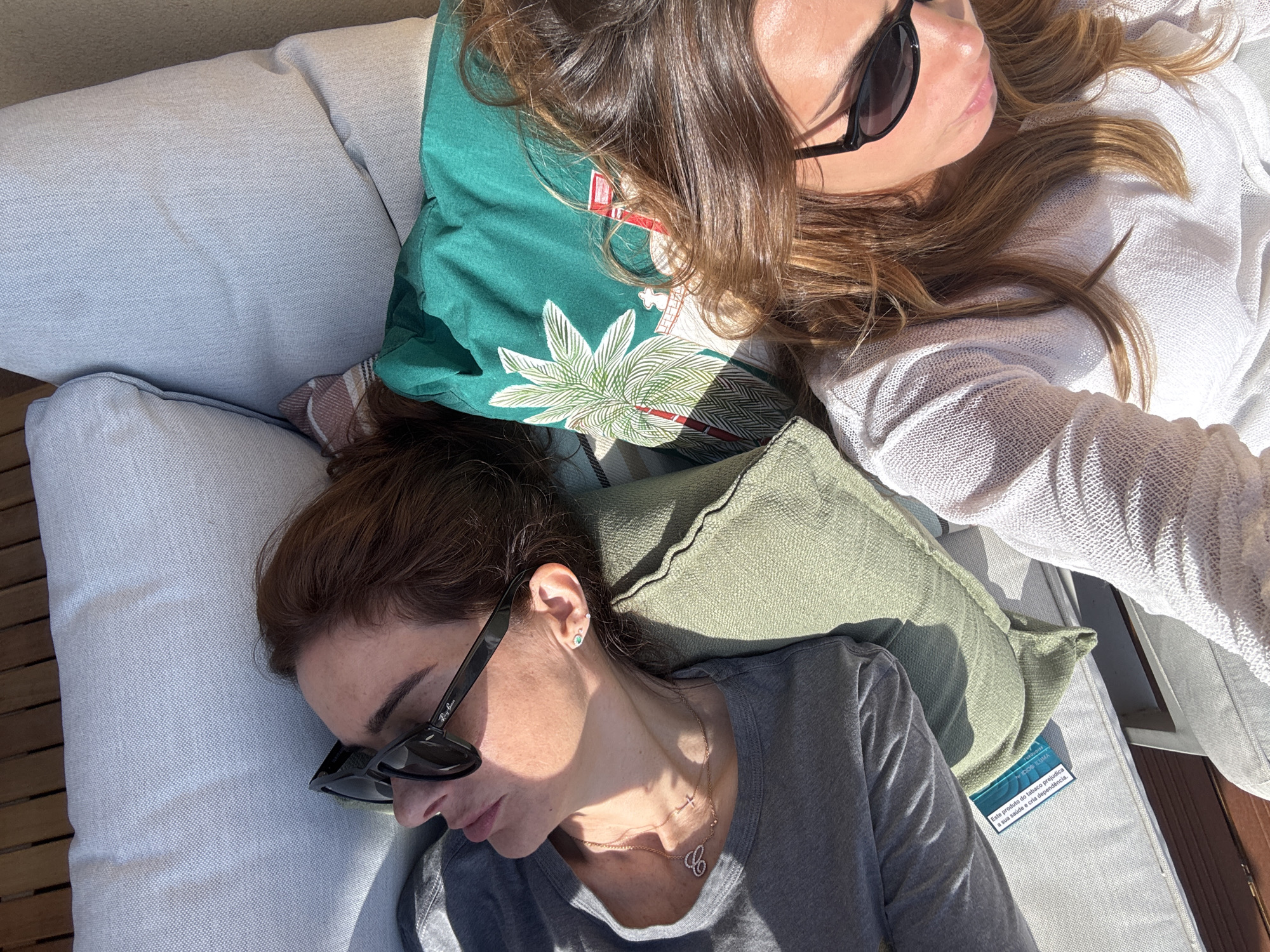 The image is an overhead shot of two women lounging on a light grey outdoor couch. One woman, wearing sunglasses and a light-colored sweater, rests her head on a green pillow printed with a tropical design. Beside her, the second woman, also wearing sunglasses and a grey t-shirt, is reclined with her head on a green pillow. The couch is adorned with several throw pillows of different colors and patterns. On the lower right corner of the image, there is a white tag that has text written in Portuguese, identifying it as an ICOS ILUMA product, which is a tobacco product. The location is Vila Nova de Gaia, Portugal.
