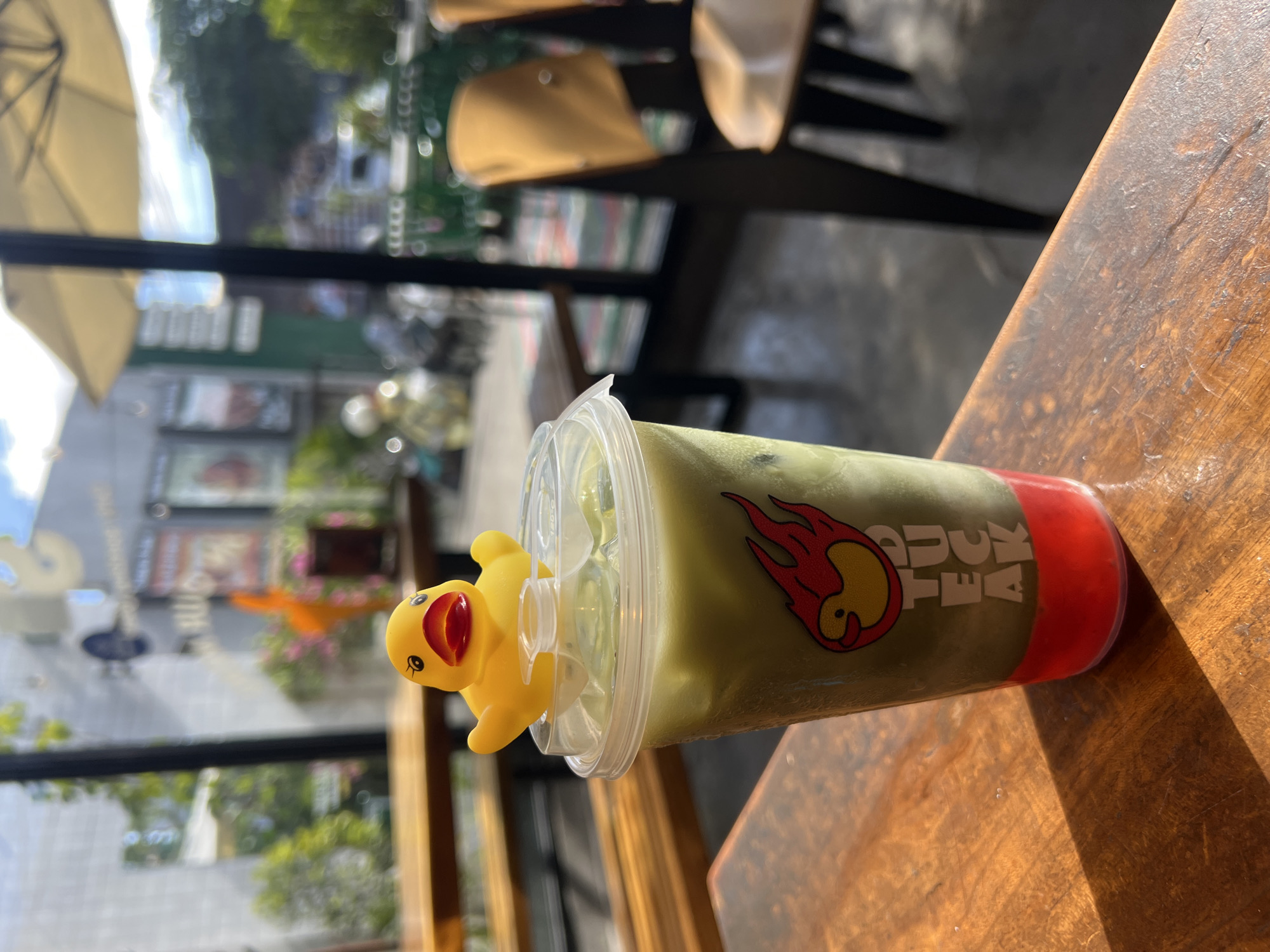 A close-up shot focuses on a plastic cup containing a green-colored beverage, likely a matcha latte, sitting on a wooden table. A small yellow rubber duck toy is perched on the rim of the cup. The cup has a logo on its side that appears to be of a duck within a fire design. The background shows a blurred street scene through what appears to be a window or open doorway. This background includes glimpses of outdoor furniture, plants, and other buildings, suggesting an outdoor cafe setting. The image was taken in the city of Kuta Selatan, Indonesia.