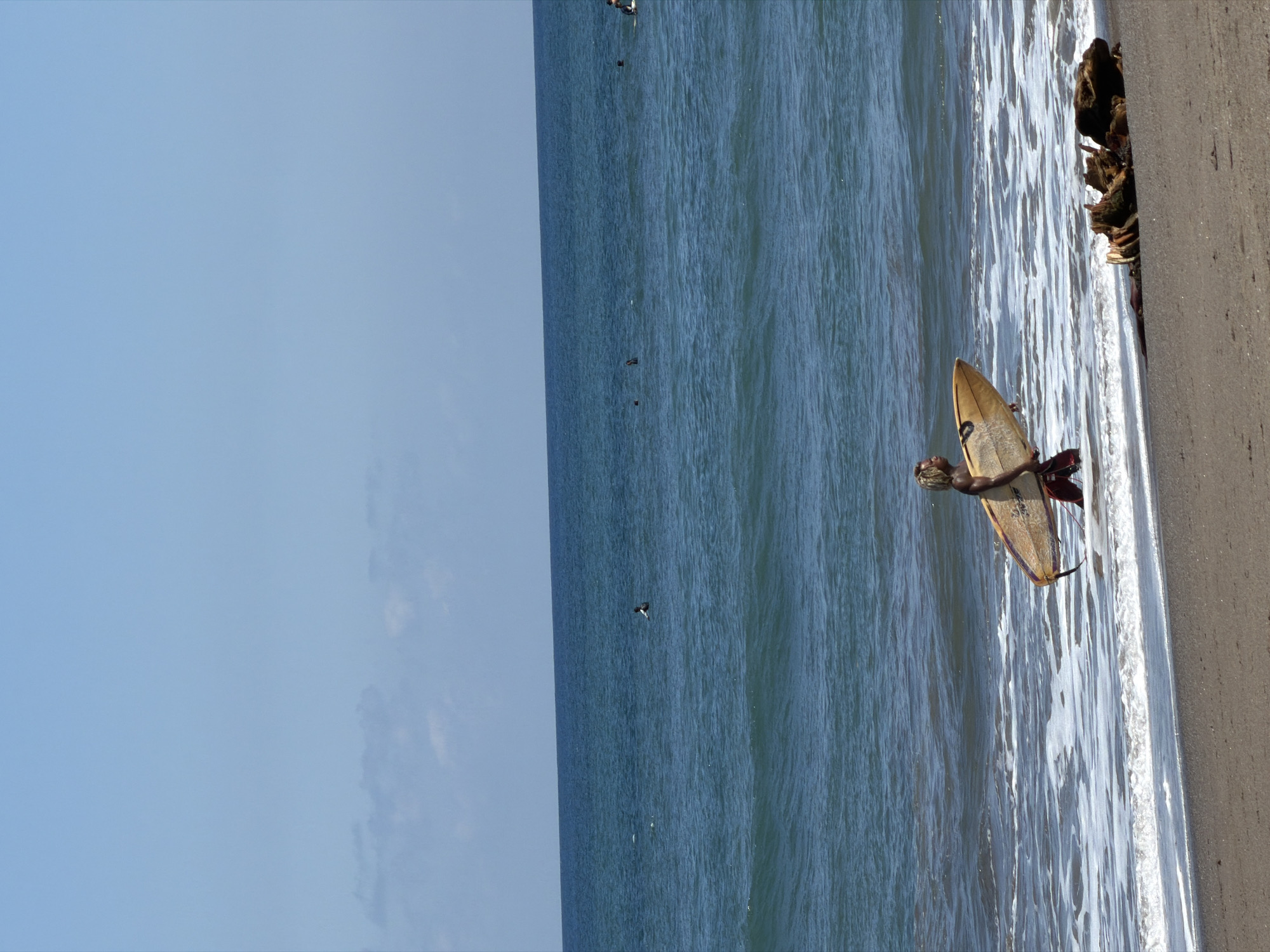 The photograph depicts a surfer at a beach in North Kuta, Indonesia. The surfer is lying on a surfboard at the edge of the water near the sand. The ocean stretches out towards the horizon, where other surfers are present. The sky is a light blue. The sand is light in color, and there are some rocks or debris near the edge of the sand.