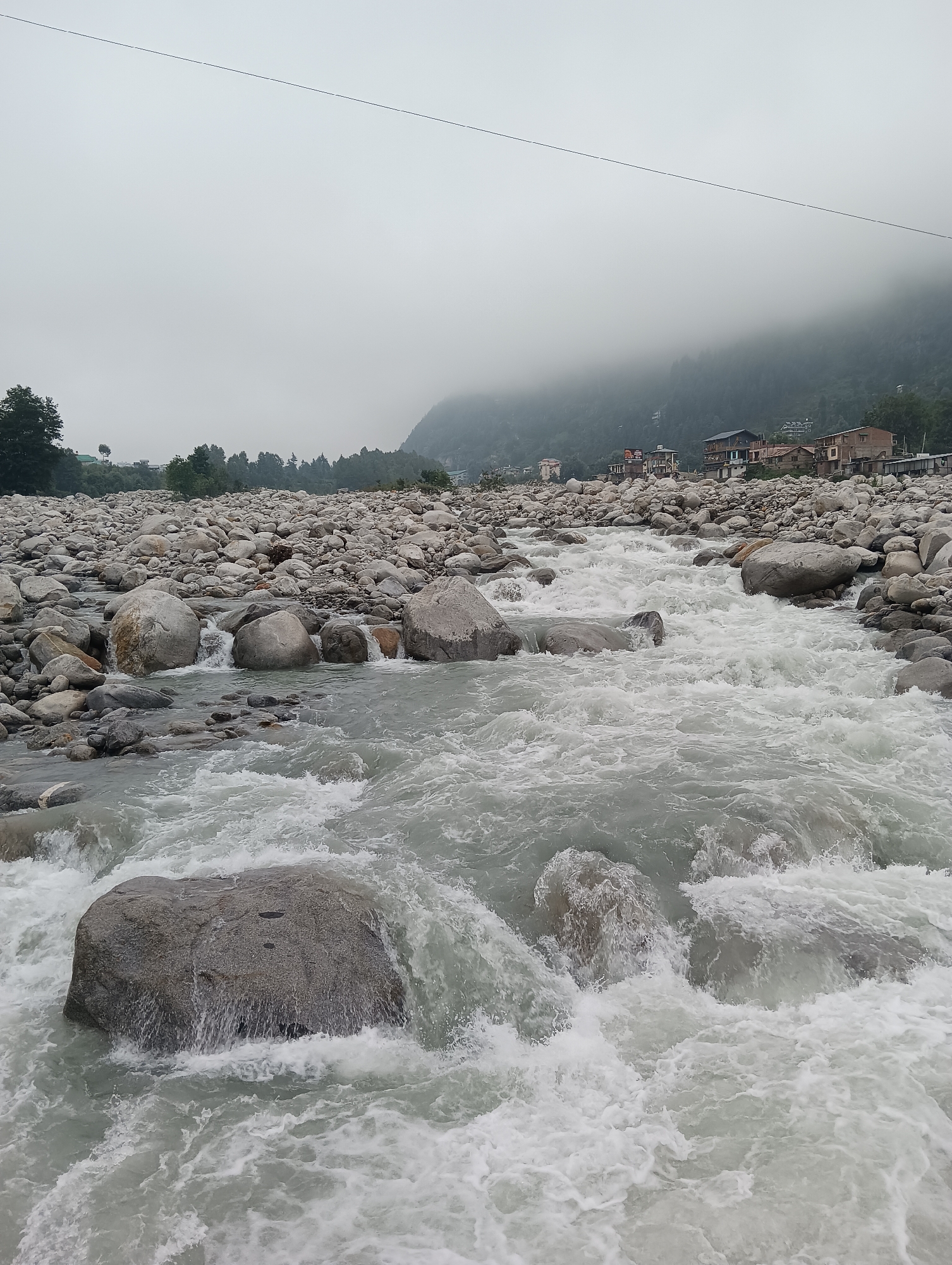 A wide, gray, turbulent river flows through a rocky, boulder-strewn riverbed in Bahang, India, under an overcast sky. The water is milky white with foam and rapids, indicating a fast current. Large, smooth, gray boulders and smaller stones litter the riverbed, with some submerged and others partially exposed. In the background, on the distant foggy hillsides, a cluster of buildings, likely homes, are visible, with trees dotting the landscape. A single, thin black power line stretches horizontally across the top of the image. The scene is desolate and wild, characterized by the raw power of nature.