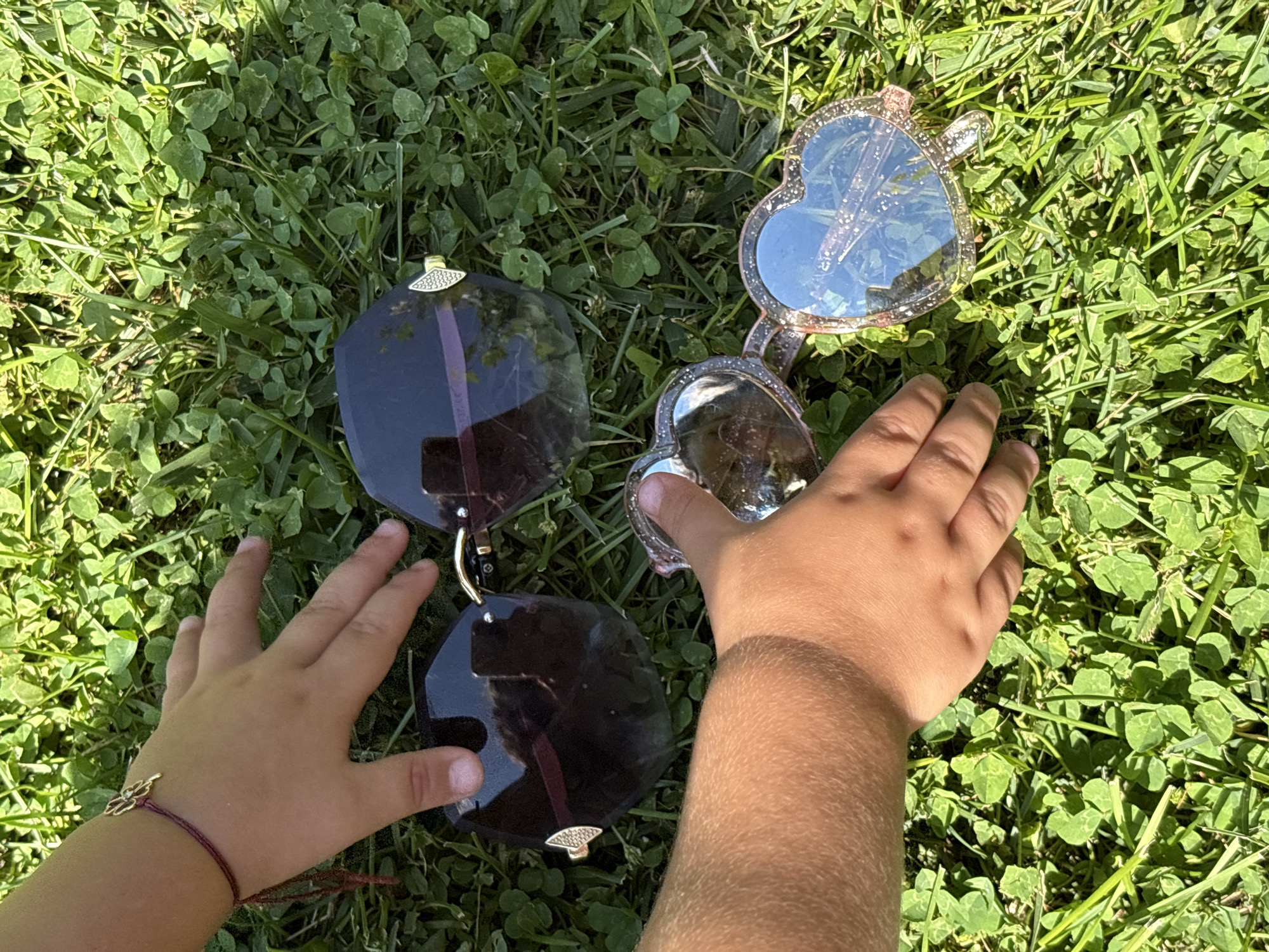 In Oreshak, Bulgaria, on a patch of sunlit green grass, two pairs of sunglasses are laid out. A child's hands are reaching for them. One pair of sunglasses is oversized with dark lenses and ornate gold detailing on the frame. The other pair is heart-shaped with a glittery pink frame and clear lenses that reflect the blue sky. A thin red string bracelet with a four-leaf clover charm is visible on the child's wrist. The image captures a moment of playful interaction with the sunglasses on a warm, grassy day.