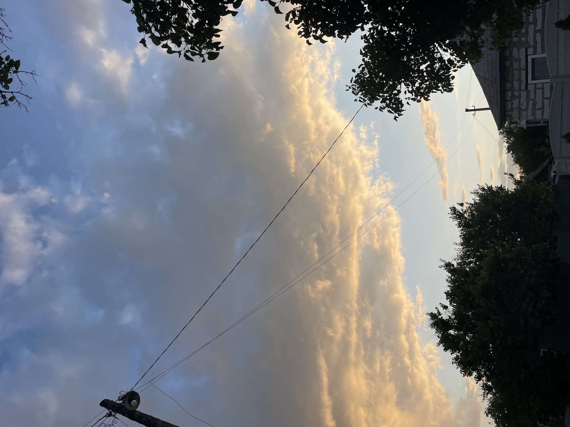 A low-angle, medium shot captures a dusky sky filled with clouds tinged with orange and yellow from the setting sun, contrasting with patches of blue. A wooden utility pole with a light fixture stands on the left, supporting taut electrical wires that stretch diagonally across the frame. Silhouetted trees frame the top right and the foreground. In the lower right quadrant, the roof of a white brick house, partially constructed and featuring a dark-trimmed window, is visible behind a white fence. The scene appears to be set in a suburban or rural area of Krasnokutsk, Ukraine.