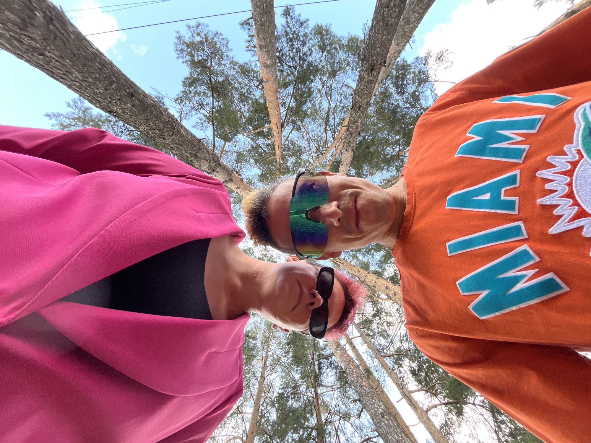 In Chaikovsky, Russia, two individuals are captured from a low-angle perspective, looking upwards towards a canopy of tall pine trees against a bright, cloudy sky. The person on the left wears a vibrant pink blazer over a dark top and black sunglasses. Their head is tilted back, and their hair is dyed pink. The person on the right wears an orange sweatshirt with large teal lettering that reads "MIAMI." They are also wearing sunglasses with a colorful, iridescent tint. Their heads are close together, suggesting they are together. The environment is outdoors, amidst a forest, with the sun filtering through the trees. The ground is not visible.