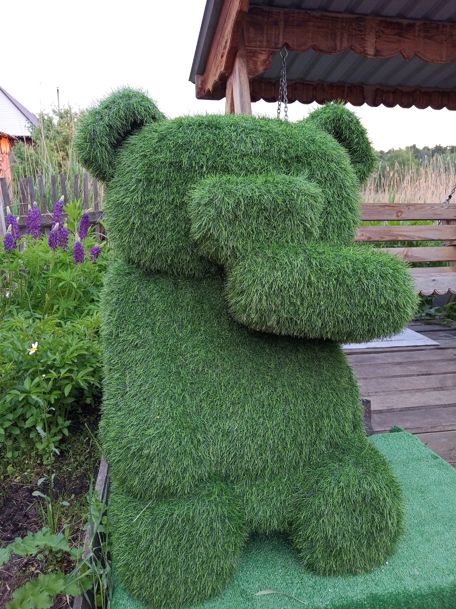 A topiary bear figure, meticulously shaped from lush green grass, stands on a green carpeted surface in the city of Pankrushikha, Russia. The bear is depicted in a seated position, with its paws raised as if covering its face. The background features a rustic wooden gazebo with a corrugated metal roof, a wooden bench, a wooden fence, and a patch of vibrant purple lupine flowers. The scene is set outdoors, with a focus on the bear as the central subject.