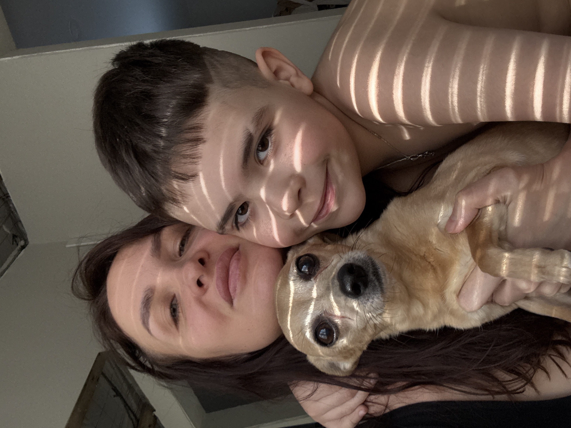 An indoor photograph captures a woman, a boy, and a small dog in close proximity, facing the camera. The adult woman, positioned on the left, has long dark hair and a subtle smile, with her left arm embracing the boy. The boy, on the right, has short dark hair with an undercut and a fringe, smiling broadly. He is wearing a thin silver-colored necklace and appears bare-chested. A small, short-haired dog with tan fur, dark eyes, and a black nose is held by the woman in the central foreground, looking directly at the camera. Strong directional sunlight enters from the left, casting distinct parallel horizontal stripe shadows across the faces and bodies of all three subjects. The background is out of focus, revealing light-colored interior walls and hints of architectural elements. The specific location within the city of Chaikovsky, Russia, cannot be determined from the visual information presented.