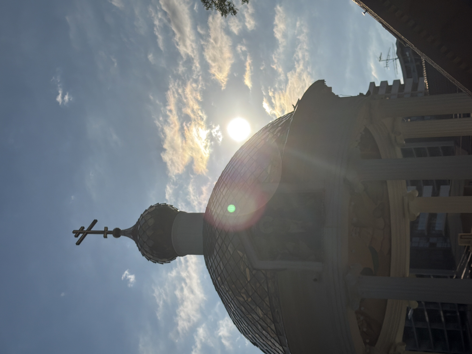 The image captures a low-angle view of an Eastern Orthodox church dome and a cross against a bright, partially cloudy sky. The sun is high in the sky, causing a lens flare that creates a green orb in the upper right portion of the dome. Wispy clouds are scattered across the sky, catching the sunlight and appearing golden. The dome itself is dark and appears to be covered in a patterned material, possibly scales or tiles. To the left of the dome, a prominent, dark Orthodox cross stands out against the sky. In the background, the silhouette of a multi-story building with a flat, crenellated top is visible, along with what appears to be a weather vane or antenna structure. The overall scene suggests a sunny day, possibly late afternoon given the angle of the light, in Sochi, Russia, a location known for its diverse architecture including Orthodox churches. No text is visible in the image. The perspective and bright sunlight create a dramatic and somewhat ethereal atmosphere.