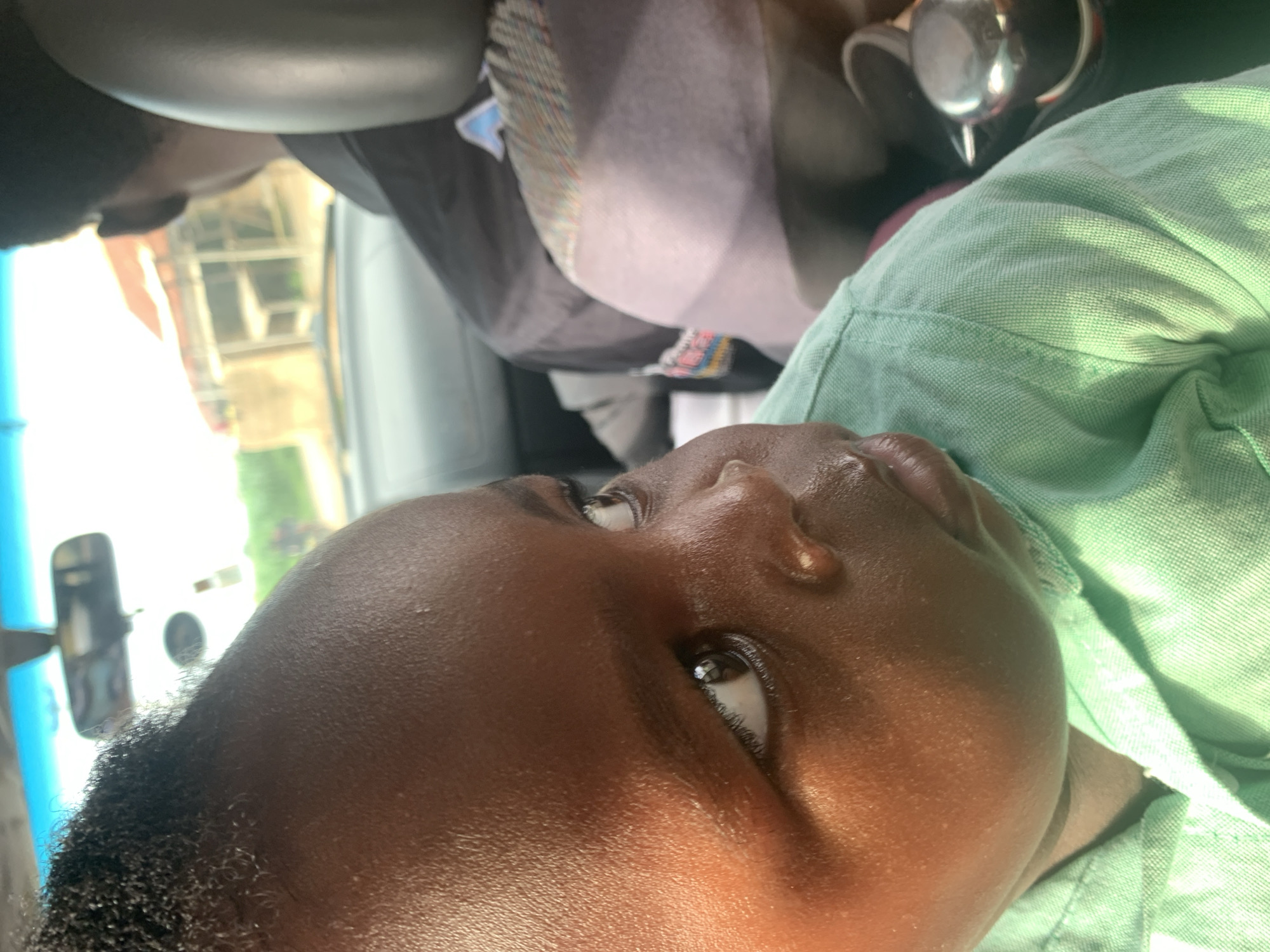 A close-up, eye-level shot shows a young child in the front passenger seat of a vehicle. The child is facing to the right, with their head tilted, and their eyes are looking outwards. Sunlight illuminates the child's face, highlighting their skin and hair. The child is wearing a light green collared shirt. In the background, out of focus, is the driver of the vehicle, wearing a dark shirt, and the interior of the car, including a rearview mirror and the dashboard. The scene suggests a moment captured during a car journey.