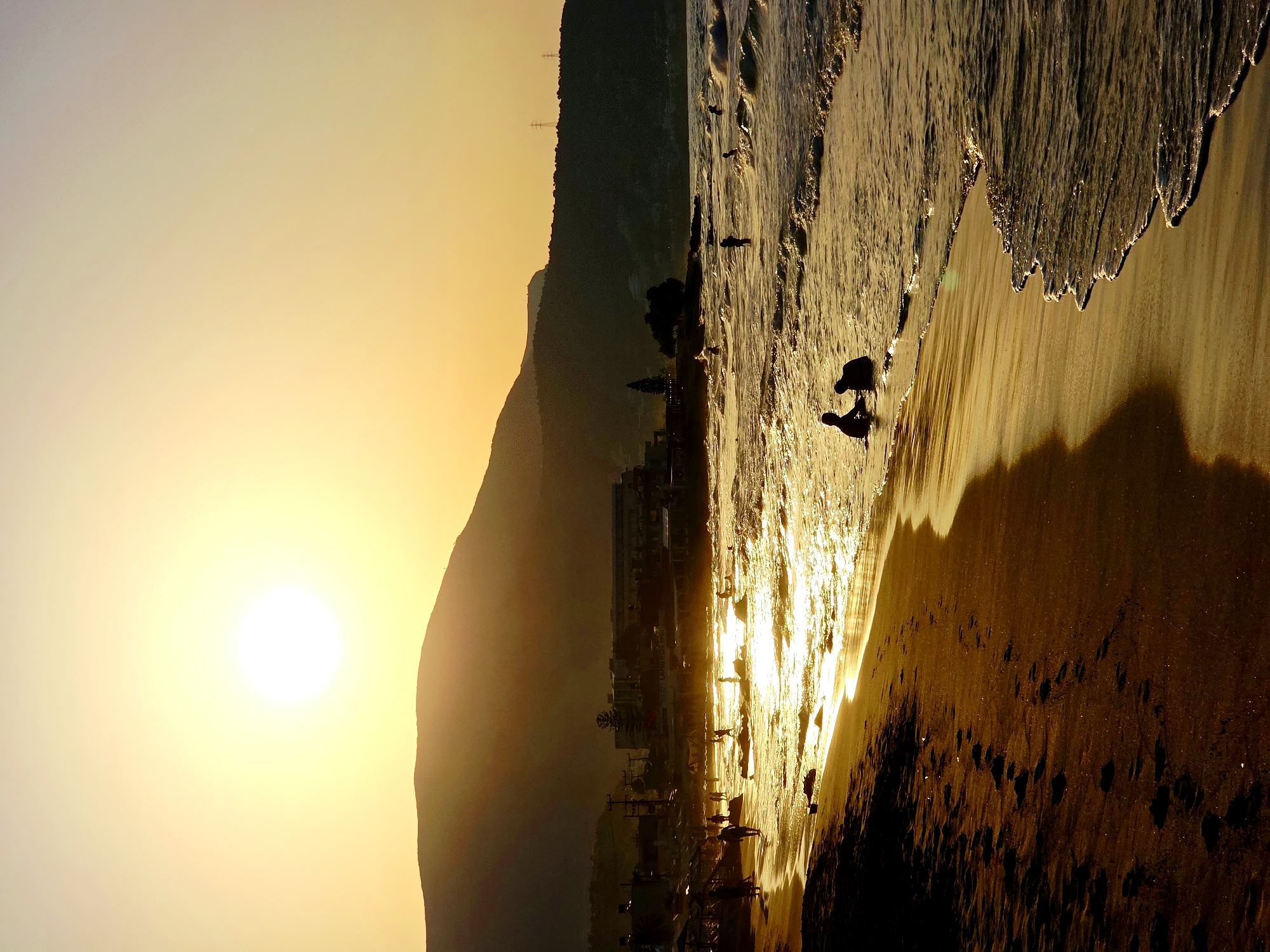 A vertical, full shot captures a sunset on the beach in Malia Municipal Unit, Greece. The sun, a bright white orb with a hazy halo, is positioned in the upper left quadrant of the frame, casting a warm, golden light across the scene. The sky transitions from bright yellow near the sun to a pale orange and then a soft lavender towards the top. Silhouetted against the sky, a large, dark mountain range forms the background, with a few buildings and structures partially visible at its base. The mountain is partly obscured by a yellowish haze from the setting sun.
The midground features the sea, with waves gently rolling onto the shore. The water is a shimmering gold due to the reflection of the sun, and white foam crests the waves. Several figures are visible in the water, appearing as dark silhouettes: two people are sitting in the shallow water in the lower right of the frame, and a few other individuals are scattered further out in the sea. The shoreline is a mix of wet sand where the waves recede and dry sand further up. Footprints are clearly visible in the dark, wet sand along the left side of the frame, leading away from the viewer. Some vegetation and structures, possibly beach umbrellas or small buildings, are visible along the edge of the beach, silhouetted against the brighter sea and sky. The overall impression is one of a peaceful, end-of-day scene on a Mediterranean beach.