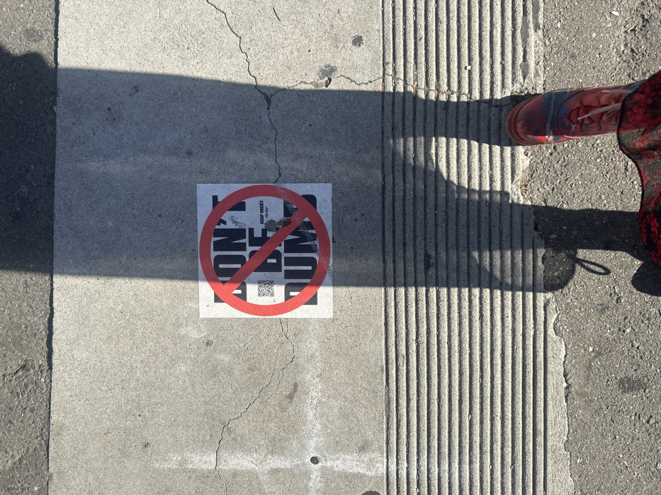 A sticker with a red prohibition symbol over the words "DON'T BE DUMB" lies on a concrete sidewalk. A QR code is also visible on the sticker. A shadow of a person's legs and feet, clad in a red and black patterned garment and a sneaker, is cast across the sidewalk and the sticker. The sidewalk has a textured edge on one side and a smooth surface where the sticker is placed. Cracks run through the concrete.