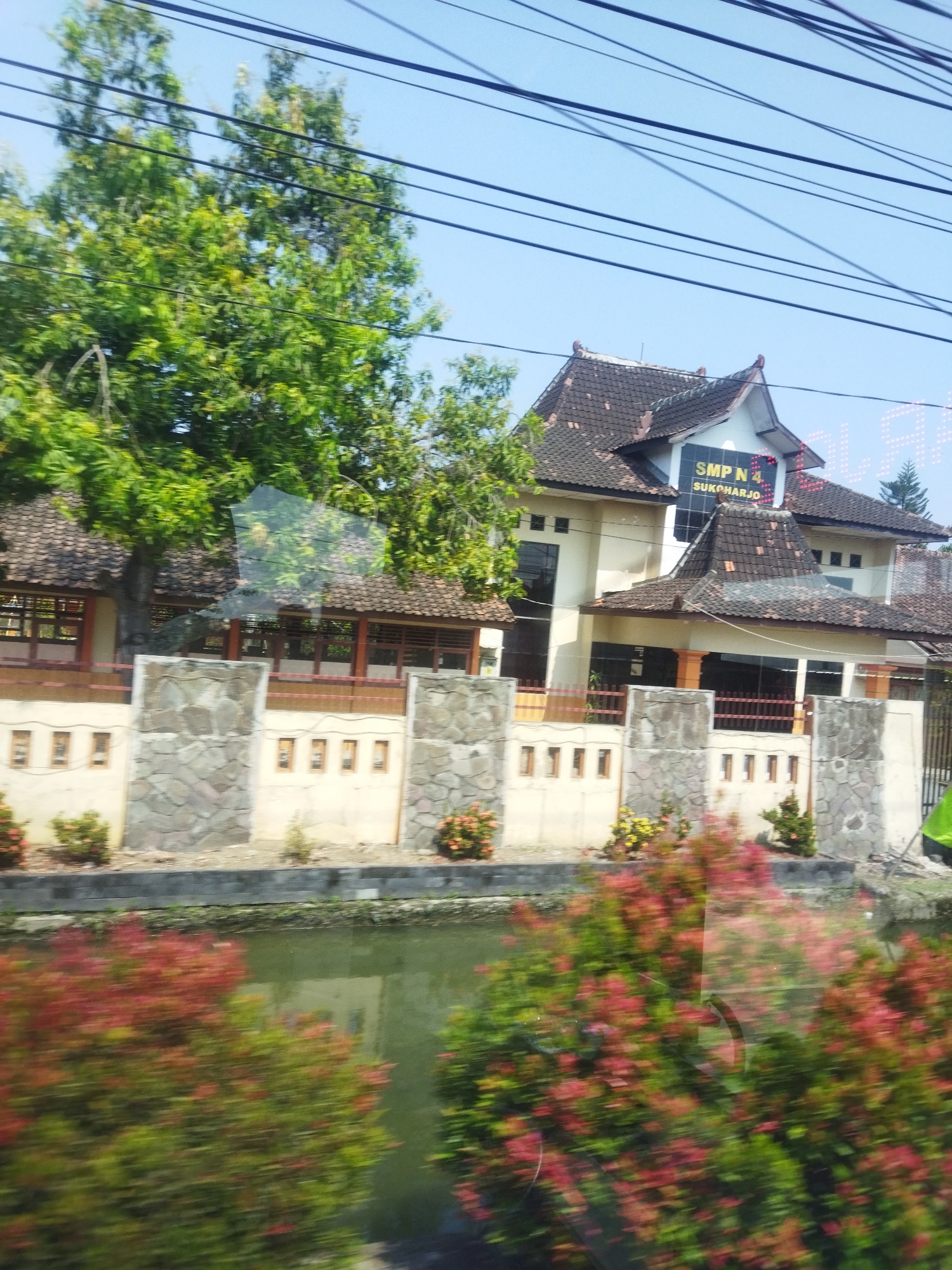 The image shows a building with traditional Indonesian architecture, a school named SMP N 4 Sukoharjo. The building has a dark tiled roof with decorative eaves and is painted in cream and orange colors. In front of the building, there is a stone wall with several small rectangular openings. Behind the wall, there is a body of water with lush green and red foliage reflected in it. The foliage appears to be on the bank of the water. The background shows a clear blue sky with several overhead power lines. The image seems to have been taken from a moving vehicle, as evidenced by the motion blur in the foreground foliage. The overall scene depicts a peaceful and somewhat rural environment within the city of Begajah, Indonesia.