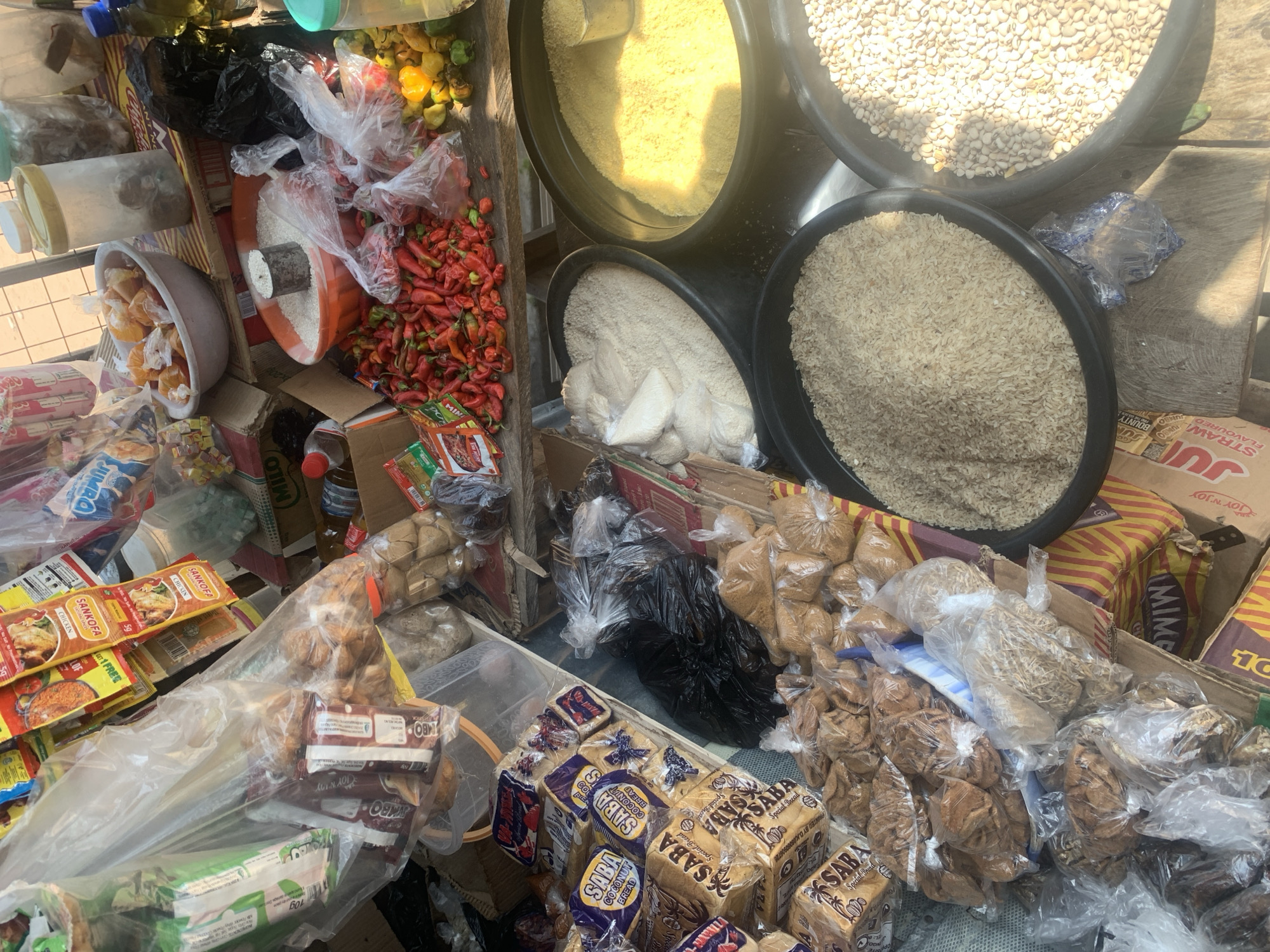 This is an overhead, close-up shot of a market stall in Jalingo, Nigeria. The stall is filled with a variety of goods, primarily food items, presented in both bulk and packaged forms.
Prominent items include large basins overflowing with grains, likely rice and possibly millet or sorghum, a common staple in Nigeria. There are also substantial piles of red chili peppers, some fresh and others dried. Smaller bowls contain what appear to be chopped oranges or mangoes. Several plastic bags hold packaged goods, including loaves of "SABA" brand bread, with varieties like "Coconut Bread" clearly visible, and smaller packets of what seem to be seasonings or spices, one labeled "SANKOFA CHICKEN". Other bagged items suggest snacks or dried goods. The overall impression is of a bustling local market where everyday foodstuffs are sold.
The lighting suggests it is daytime, with sunlight casting some shadows. The setting is outdoors, likely a street market, indicated by the rough wooden surfaces and the general arrangement of goods. There are no people prominently featured in the frame, focusing solely on the merchandise. The visible text on packaging, such as "JOY 'N JOY", "JUMBO", "BOUNTY", "MILO", and the "SABA" bread labels, provides specific details about the products and their brands. The scene conveys the abundance and variety typical of an African market.