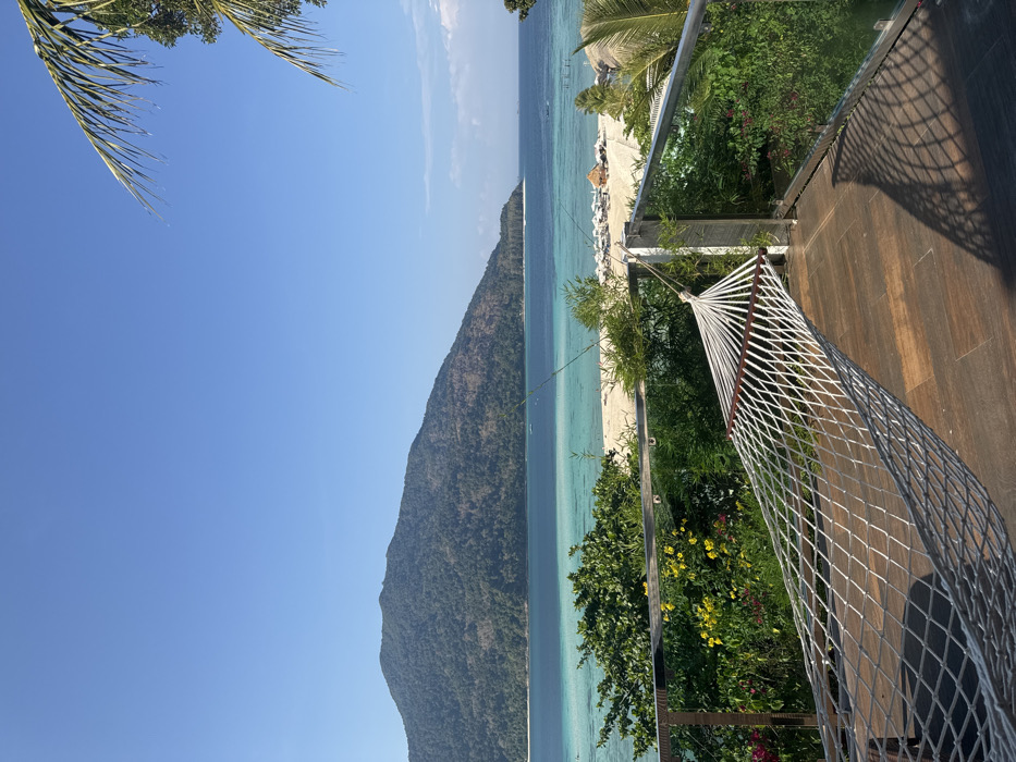 The image presents an elevated, panoramic view of a tropical coastal environment in Ban Ko Li Pe, Thailand, under a clear blue sky. In the immediate foreground, a white, rope-net hammock with wooden spreader bars is suspended on a dark wood-paneled balcony. The balcony is bordered by a railing constructed of stainless steel posts and glass panels. Sunlight, originating from the right, casts distinct shadows of the hammock netting and railing onto the balcony floor. Lush green and yellow flowering foliage is visible just beyond the railing.
The midground features a curved white sand beach, bordered by a calm sea exhibiting distinct bands of turquoise in the shallows and deeper blue further offshore. Numerous white beach umbrellas and sun loungers are arranged on the sand, alongside a few small, thatched-roof structures. Several small boats are visible in the water. A large, heavily forested island or mountain dominates the background, stretching across the horizon. A portion of a palm frond is visible in the upper right quadrant of the image. No individuals are present within the frame.