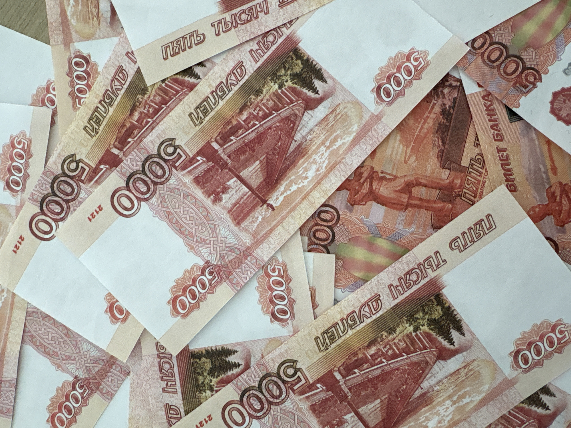 The image shows a pile of Russian banknotes with a face value of 5000 rubles. The notes are piled irregularly and partially overlap each other. The banknotes feature a design with depictions of the city of Yembaevo, Russia. Key details visible on the banknotes include the denomination "5000", the words "Пять тысяч" (Five Thousand), and images of a bridge and monument which are landmarks of the city. The image appears to be a close-up.