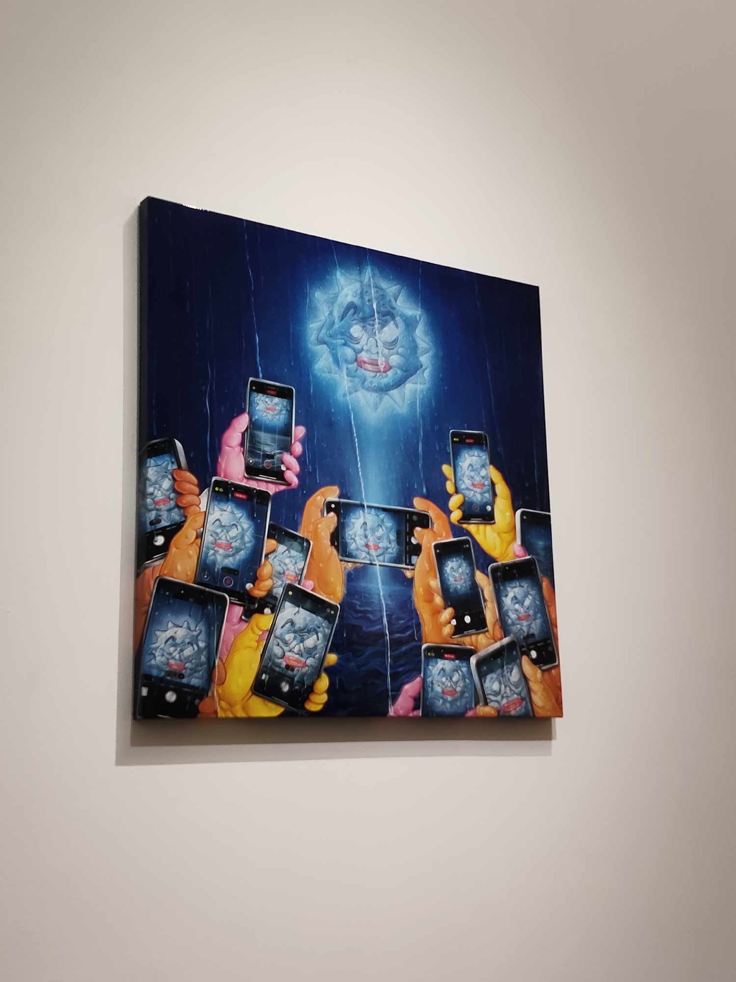 A framed artwork, likely a canvas print, hangs on a plain white wall. The artwork depicts a surreal scene with multiple hands, in various shades of yellow and orange, holding up smartphones. Each smartphone displays a cartoonish, spiky blue creature with red lips and a wide, surprised mouth. The creature appears to be illuminated by a bright light originating from above, which also casts a blue glow on the background, suggestive of rain or water. The overall aesthetic is vibrant and somewhat unsettling, with the repetition of the creature and the phones creating a sense of mass consumption or fascination. The perspective of the photograph is from a slightly low angle, looking up at the artwork.