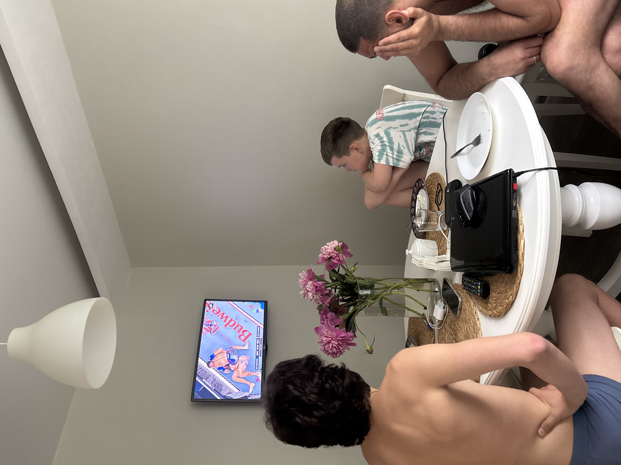 The image depicts an indoor scene, possibly a dining room or kitchen area, with three individuals seated around a white circular table. The room has light grey walls and a white ceiling with a prominent white pendant light fixture.
On the wall above the table, a flat-screen television displays a mixed martial arts fight, with a "Budweiser" logo visible on the canvas. In the foreground, a young man with dark, curly hair is seated with his back to the camera, facing the television. He is shirtless and wearing blue boxer briefs. To his right, a young boy in a tie-dye t-shirt is also seated and facing the television, with his arms crossed. To the far right, another man, shirtless and wearing dark shorts, is seated with his head in his hand, looking down. His pose suggests discomfort or weariness.
On the table, there are several items including a vase of pink peonies, a sugar bowl, plates with leftover food, glasses, a mobile phone, a remote control, a laptop, and a fork. The presence of the laptop and phone suggests the individuals might be engaged in or have recently finished some activities other than just watching television. The overall atmosphere appears casual and relaxed. There is no indication of the city of Chaikovsky, Russia.