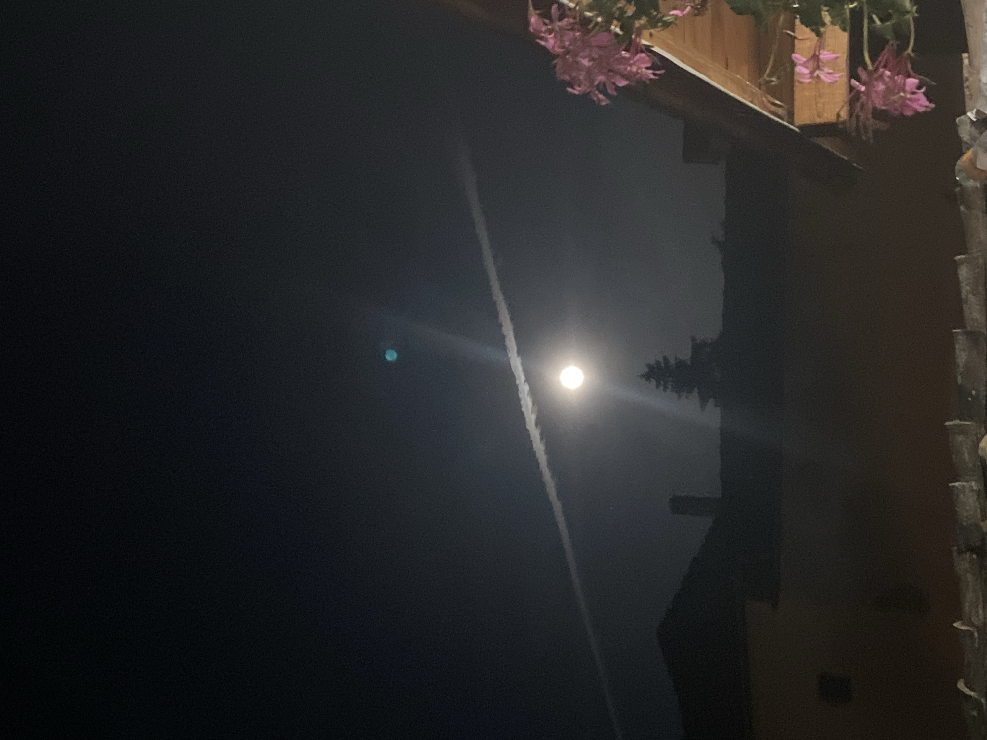 The image, taken at night, depicts a bright moon with a visible contrail extending from it in a dark sky. To the right of the sky are what appear to be the dark outlines of a building, with a flower box above it. The flowers are pink. The image may have been taken in Mindia, Bulgaria. The photo may have been rotated sideways.