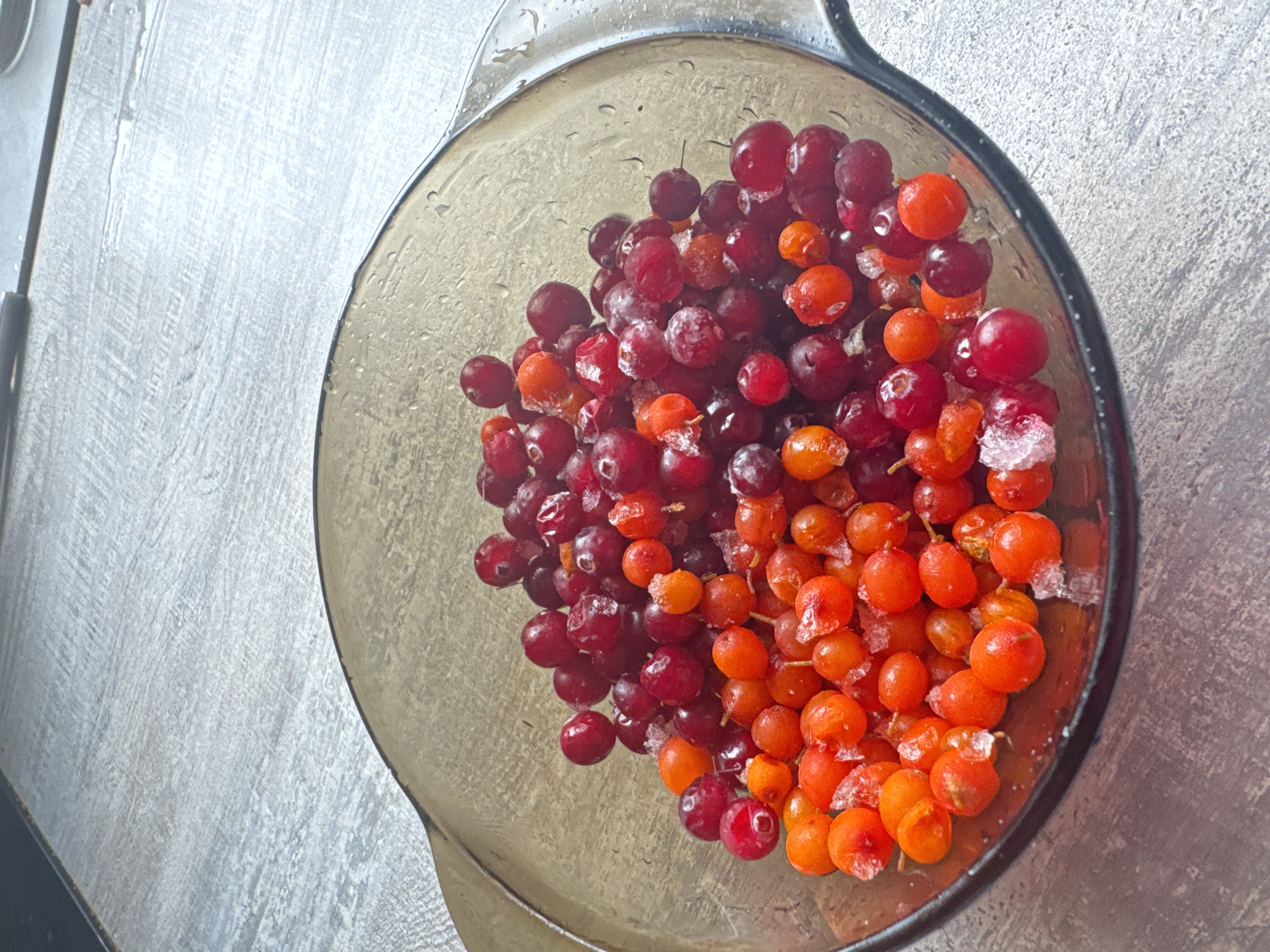 A high-angle shot captures a translucent, dark-tinted bowl filled with a mix of small, round berries. The berries are primarily of two colors: dark red/burgundy and bright orange. Many of the berries, particularly the orange ones, are covered with a visible layer of frost or ice crystals, indicating they are frozen or have recently been removed from a freezer. Water droplets are visible on the interior and exterior of the bowl, as well as on some of the berries, suggesting condensation or thawing.
The dark red berries are concentrated more towards the left side of the bowl, while the bright orange berries dominate the right side, creating a clear visual division. The background is a textured, light-colored surface, possibly a countertop or table with a wood-grain or stone-like pattern, appearing in shades of light gray and white. A dark edge of what might be another object or part of the bowl's handle is visible on the far left.
The scene depicts these berries, likely cranberries or lingonberries (red) and sea buckthorn or a similar berry (orange), resting in the bowl. No specific activity is occurring, but the frosty appearance suggests they are being thawed or prepared for cooking/consumption. The setting is an indoor environment, consistent with a kitchen. There are no people or visible text in the image. The provided location context is Chaikovsky, Russia.