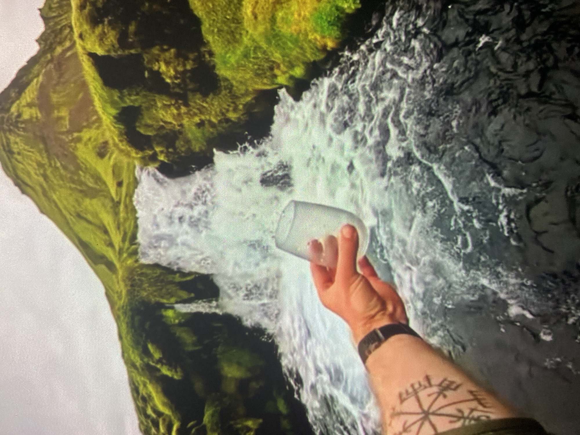 A person's arm and hand are visible in the foreground, holding a clear glass to the right. The arm has a dark tattoo on the forearm. The background is a scenic landscape featuring a waterfall cascading down moss-covered rocks and a vibrant green mountain. The body of water in the foreground is dark and turbulent. There is no indication that this scene is located in the city of Ustyvytsia, Ukraine.