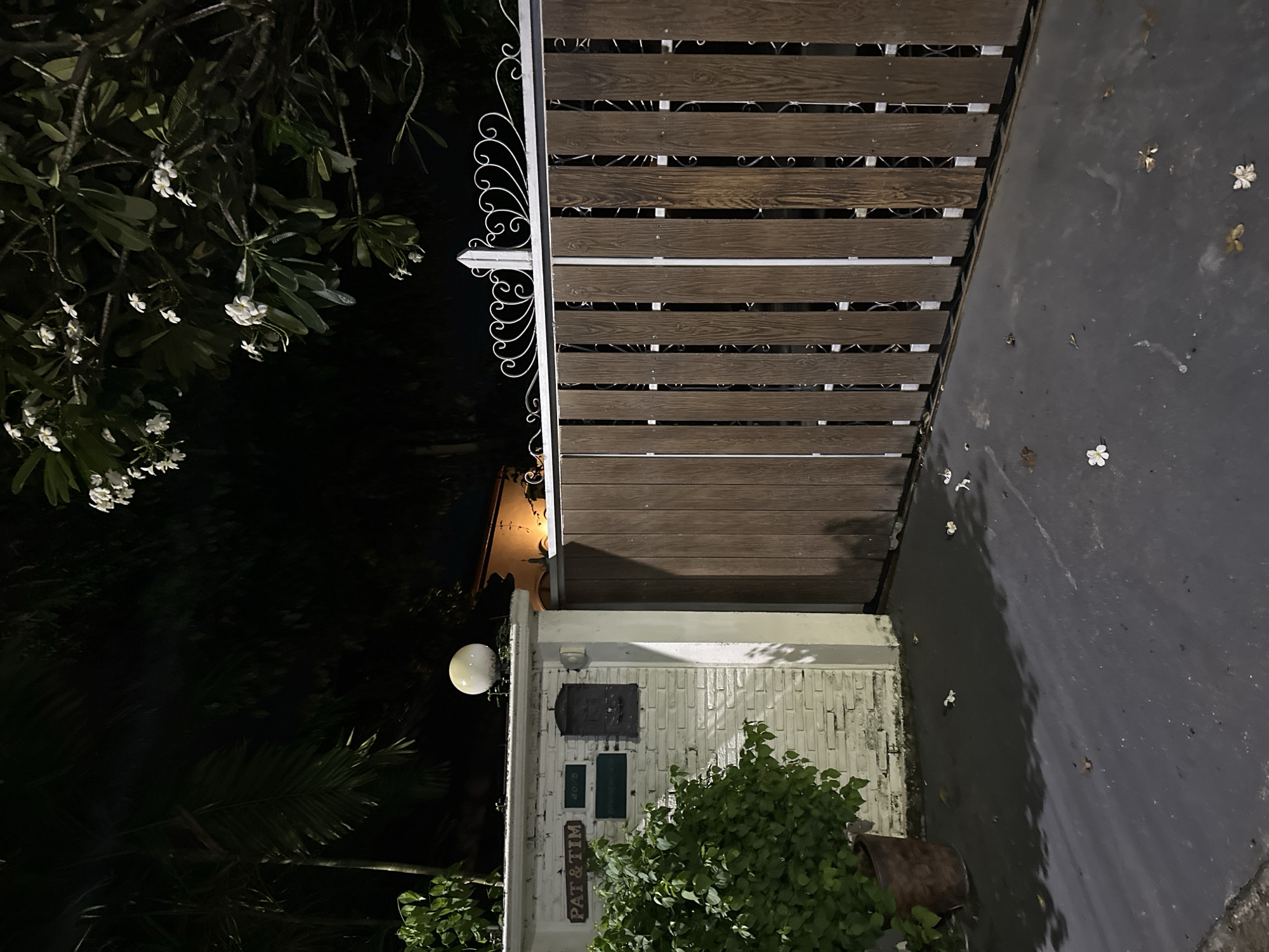 This is a nighttime, eye-level, outdoor shot of a gate and a portion of a wall in an urban setting. The gate is made of vertical wooden planks with decorative metalwork at the top. To the left of the gate is a white brick pillar that has a sign that reads "PAT & TIM", a mailbox, and a spherical light fixture on top. A large green bush in a brown pot is situated next to the pillar. The ground is paved and shows some scattered white flower petals. The background is dark, with some foliage and a building with a lit window visible.