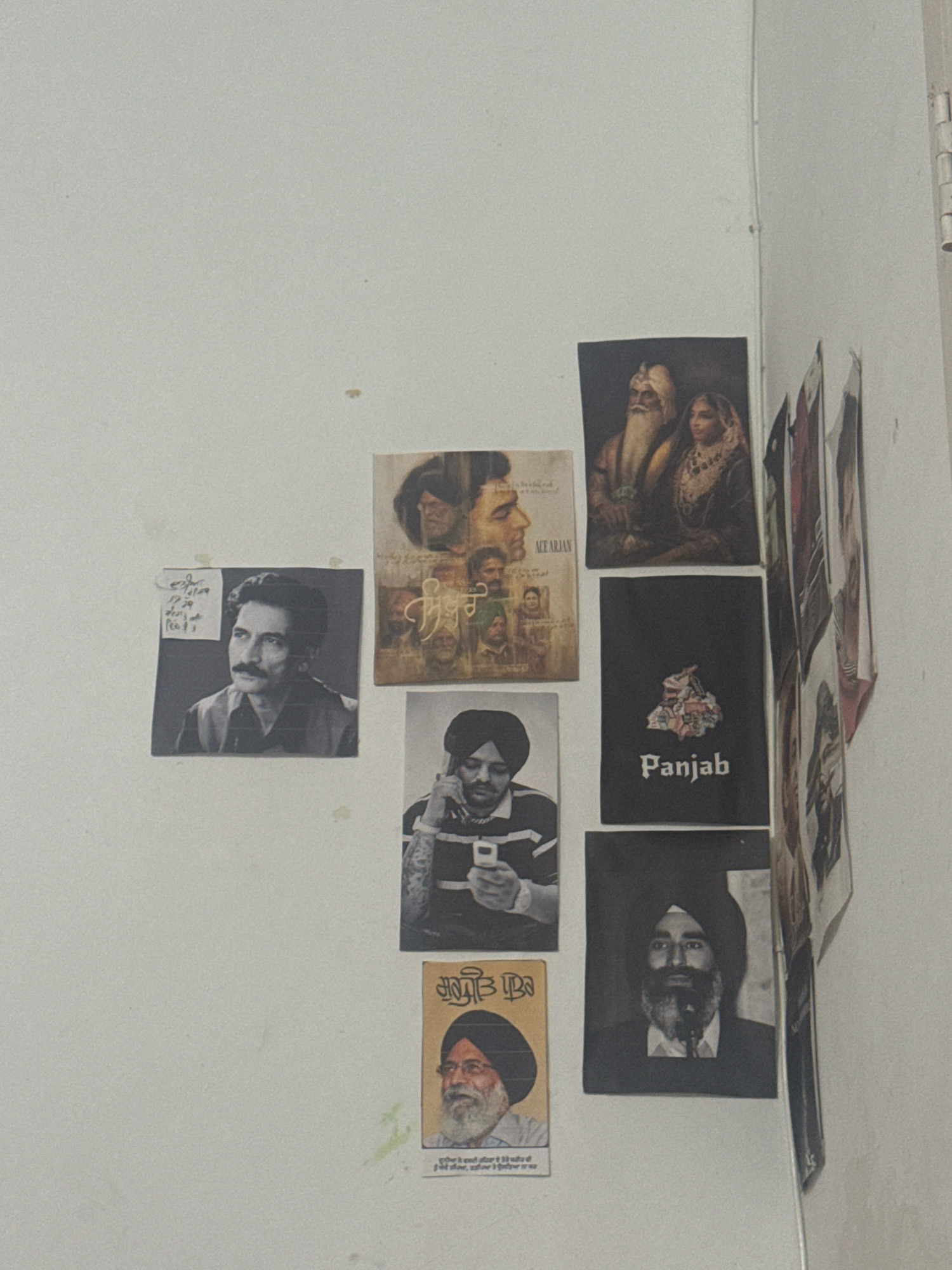 The image displays a collection of posters and photographs adhered to a white, textured wall, likely in a residential setting in Wandar Jatana, India. The collection features several portraits and graphic designs.
On the left, a black and white photograph shows a man with a mustache looking to the right, with some handwritten text in Punjabi on a smaller paper attached to its left. Above this, a framed poster is partially visible, depicting a montage of faces and text in Punjabi.
In the center, a poster features a portrait of a man in a turban holding a mobile phone. Below this is another poster with Punjabi text and a portrait of an elderly man with a beard and turban. To the right of the man with the phone, a poster displays a stylized map of Punjab with the word "Panjab" written below it in a gothic font.
Above the "Panjab" poster, a colorful poster shows a portrait of an elderly man with a long beard and a woman in traditional attire.
To the far right, several other posters and photographs are partially visible, leaning against the wall or partially overlapping, some appearing to have religious or cultural imagery. The wall exhibits some minor blemishes and marks. The overall scene suggests a personal display of cultural and artistic interests.