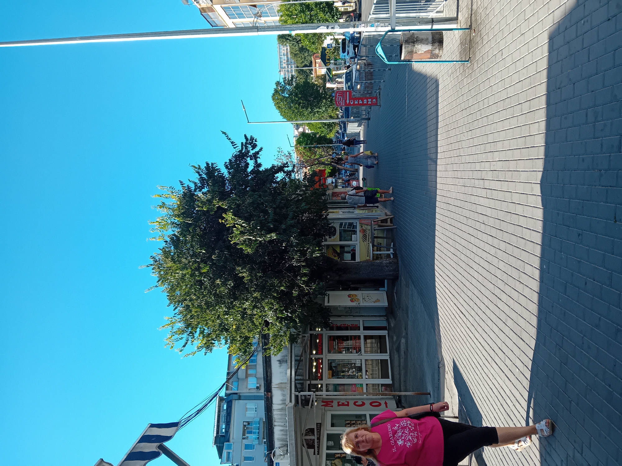 The image depicts a sunny street scene in Varna, Bulgaria, with a clear blue sky overhead. A paved sidewalk runs alongside a row of shops and buildings. In the foreground, a woman wearing a pink t-shirt and black leggings walks on the sidewalk, casting a long shadow. A large tree stands in the middle of the scene, providing shade. The shops appear to be small businesses, with storefronts displaying various goods and signage. Further down the street, more trees and buildings are visible, as well as a street with cars. A metal trash can with a blue frame is situated on the right side of the sidewalk. The overall impression is that of a typical day in a bustling urban environment.