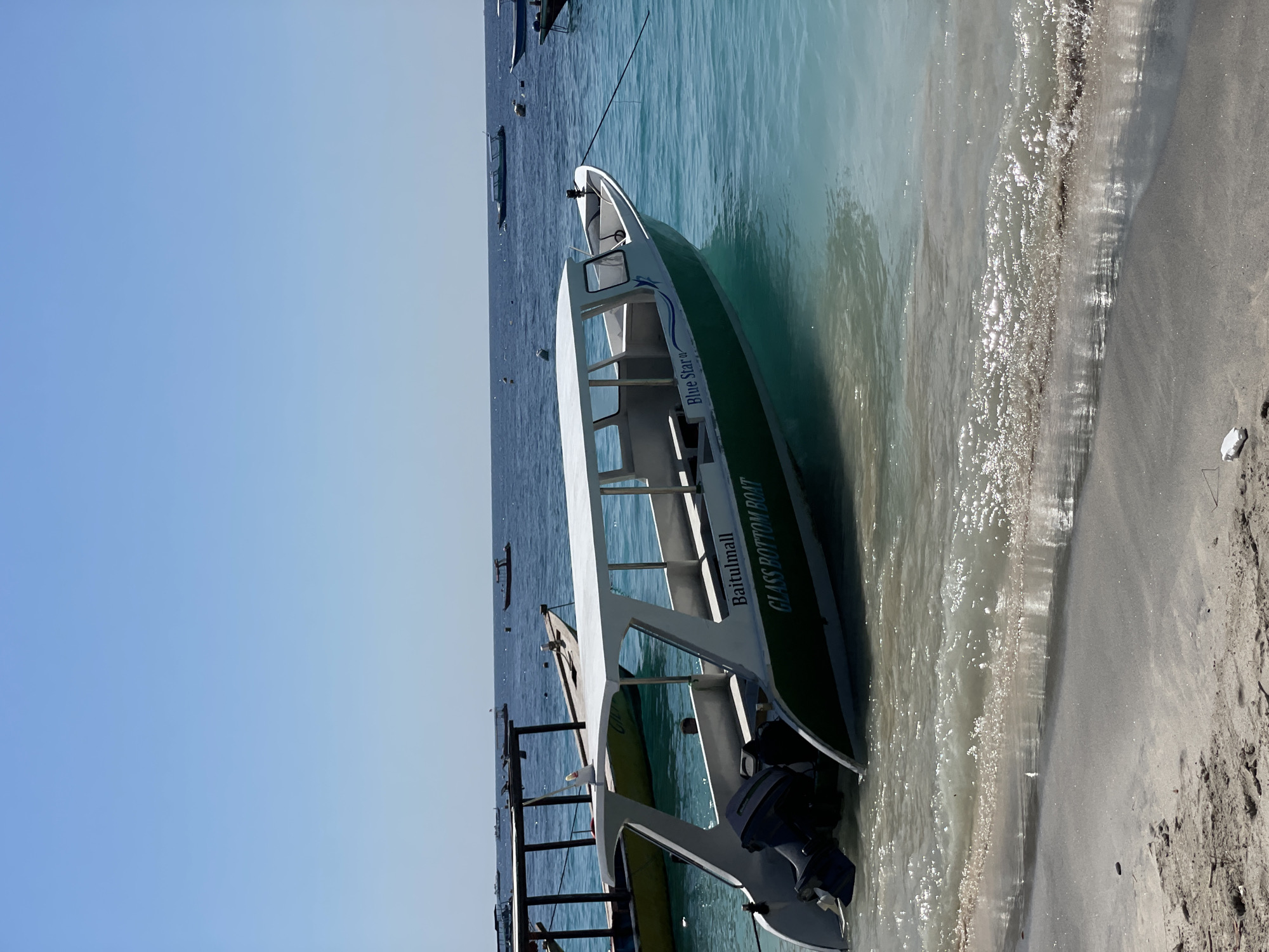 A glass bottom boat named "Blue Star" is moored in shallow, turquoise water near a sandy beach. The boat is white with green trim and has an outboard motor at the stern. Several other boats are visible in the background, as well as a wooden pier structure. The sky is clear and blue. The scene appears to be a tourist destination, likely in Indonesia, given the type of boats and the environment.