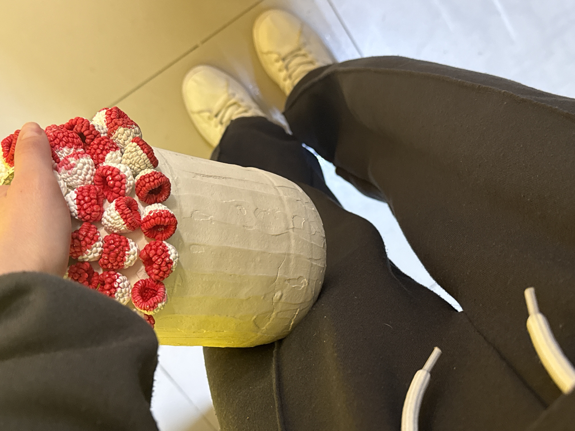 The image presents a close-up, elevated view of a person sitting indoors, showcasing their lap and a handcrafted object. The person is wearing dark gray or black sweatpants and off-white sneakers. In their left hand, they hold a cylindrical, handmade item decorated with red and white raspberry-like embellishments on top. The base of the object appears to be painted with white and yellow. The floor is a light neutral tile. The location is described as Unknown, Unknown.