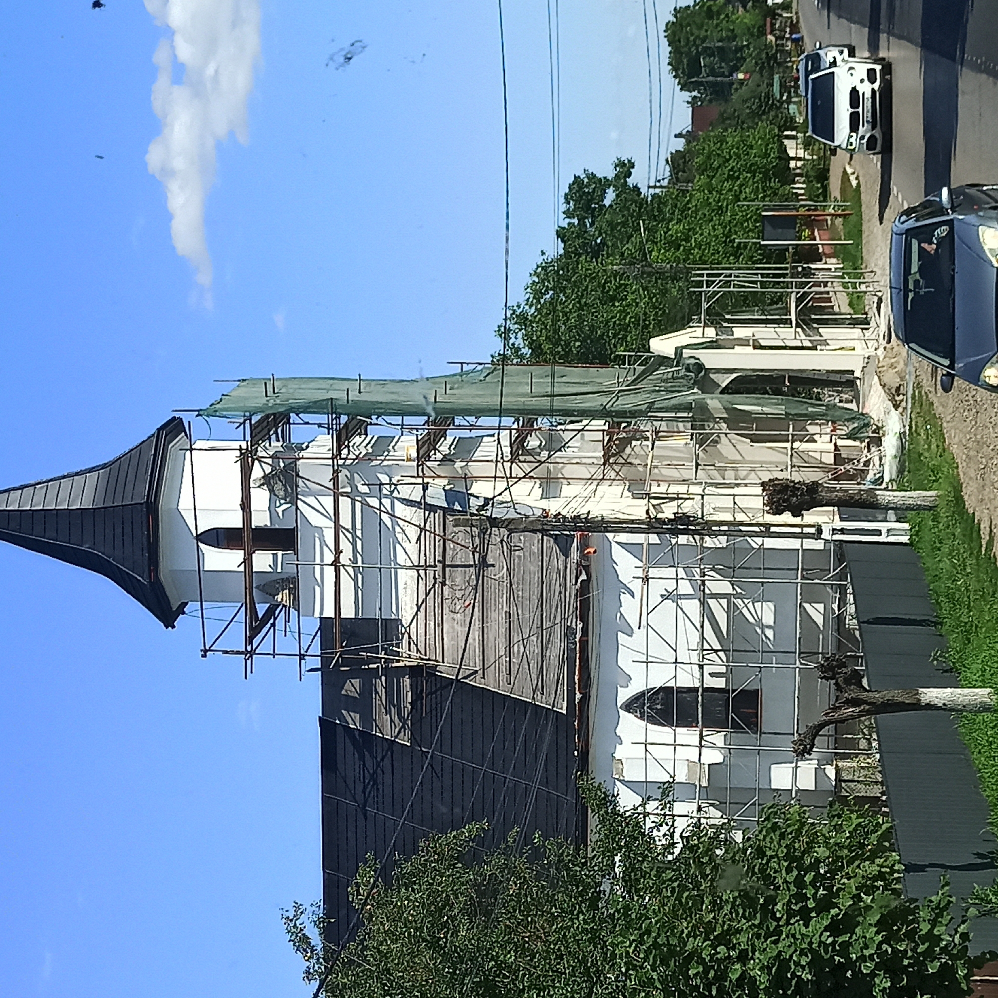 The image depicts a church under renovation in the city of Milișăuți, Romania. The church, primarily white with a black, pointed spire, is heavily covered in scaffolding and green netting. The roof appears to be undergoing repair or replacement. To the left, a large, leafy green tree partially obscures the church. In the foreground, a gray car is parked on a gravel road. Beyond the church, a street lined with trees and houses is visible, with several cars driving on the road. The sky is clear blue with a few scattered clouds. The overall impression is one of ongoing construction and daily life in a Romanian town.