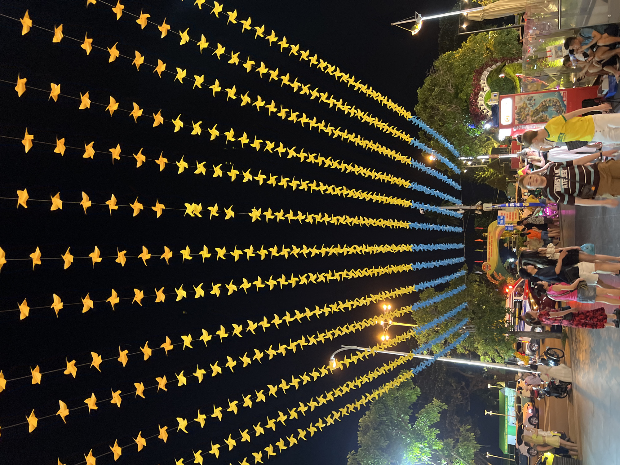 The image captures a vibrant night scene in Nha Trang, Vietnam, likely a pedestrian area or marketplace. Overhead, strings of yellow and blue paper windmills or pinwheels are suspended against the dark night sky, creating a festive canopy. The windmills are arranged in multiple rows, extending across the width of the frame.
In the foreground and midground, a crowd of people is visible. They are engaged in various activities: walking, standing, and interacting. Several individuals are the focus, including a man in a striped t-shirt and shorts, a man in a yellow t-shirt and white shorts, and groups of women standing together. Some people are dressed casually for a warm evening.
The environment includes streetlights casting a warm glow, trees with lush foliage, and what appears to be the entrance to an amusement or entertainment venue with an arched structure decorated with colorful elements. Various vehicles, including cars, a motorcycle, and potentially an ambulance or police car with flashing lights, are present on the side. The overall atmosphere suggests a lively public space at night, possibly during a festival or a busy commercial period.