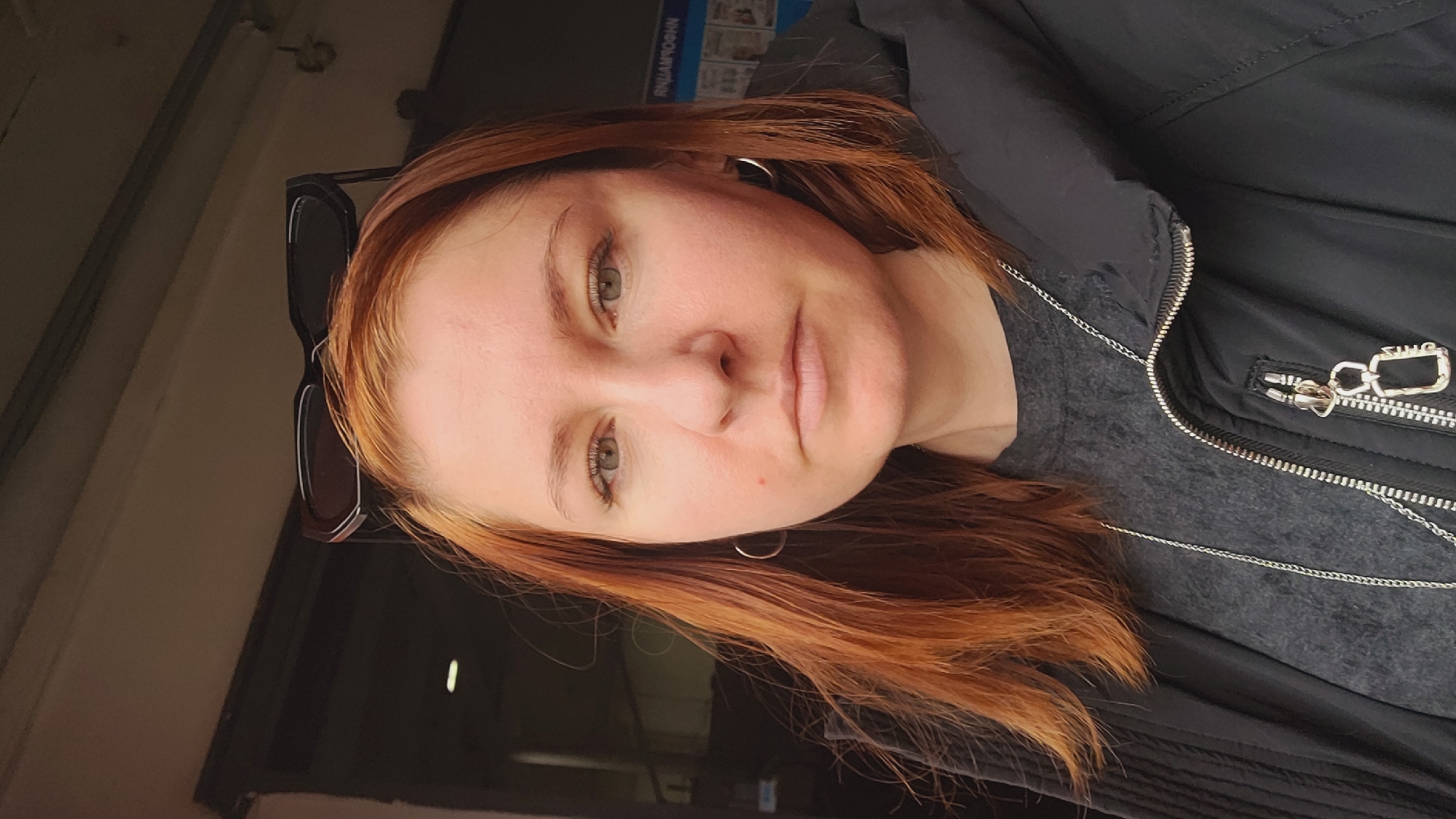 A person with red hair and a grey t-shirt is wearing a black jacket with a silver zipper. The person has sunglasses on their head and is wearing silver hoop earrings. The location appears to be Unknown, Unknown.