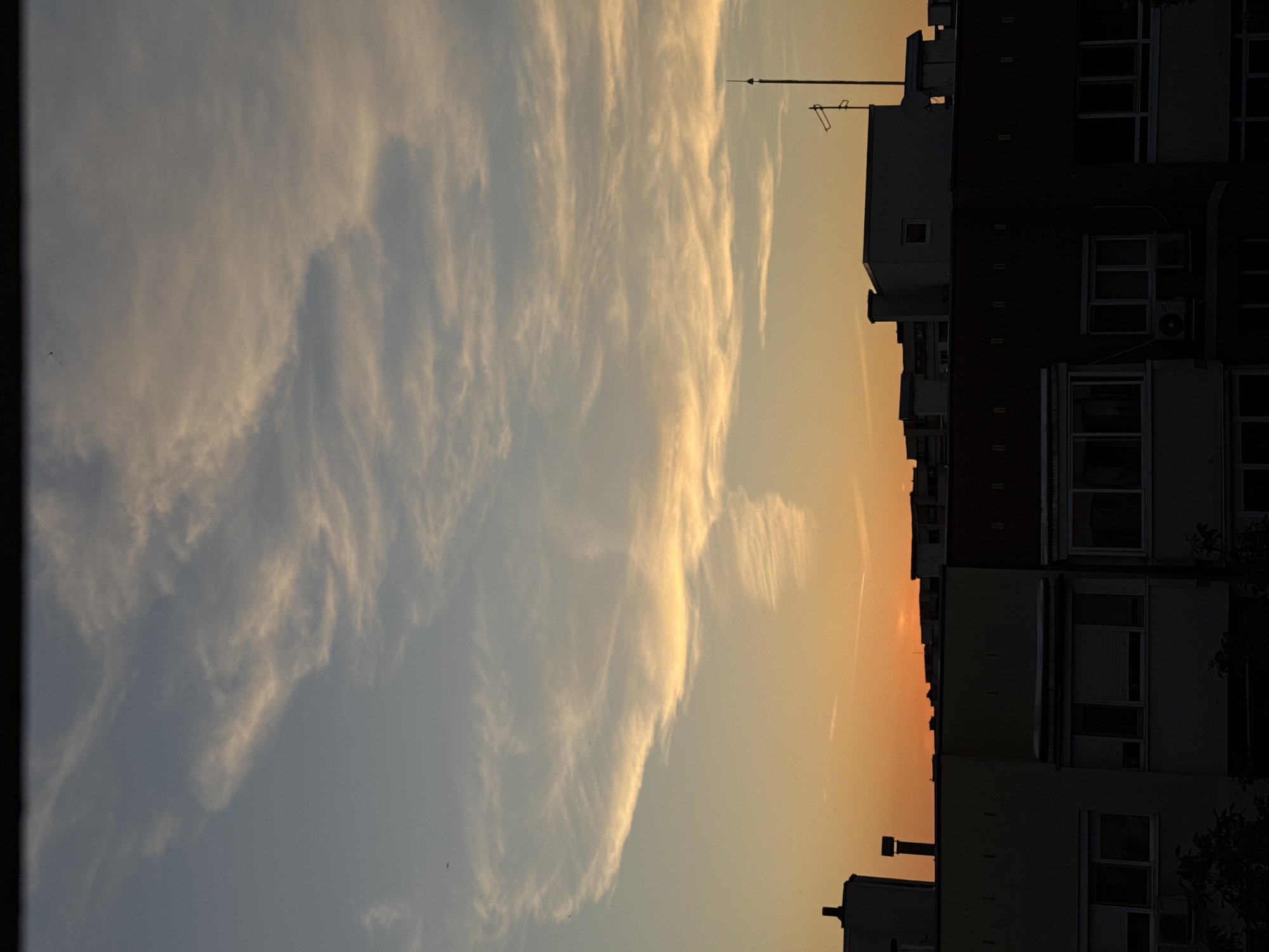 A view of the sky above Pleven, Bulgaria at sunset. The sky transitions from a pale blue at the top to a warm orange near the horizon, filled with wispy, golden clouds. Silhouetted against the sky are the rooftops of apartment buildings. These buildings exhibit varied architectural styles, with some featuring chimneys and antennae. The lower portion of the image shows the darkened facades of the buildings, with glimpses of windows and balconies. The overall impression is a calm, urban evening scene.