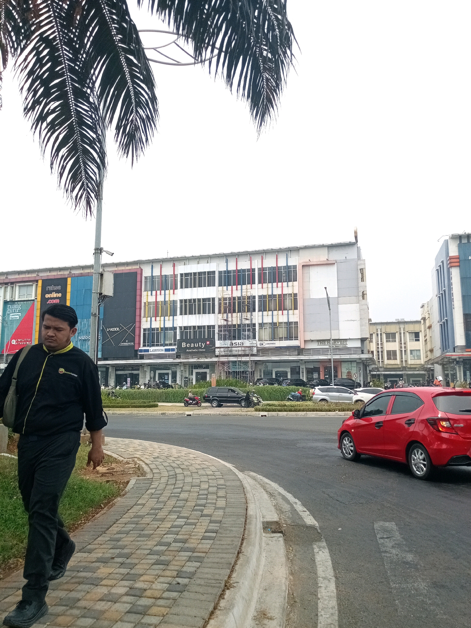The image depicts a street scene in Bekasi, Indonesia. In the foreground, a man wearing a black jacket with a yellow collar and black pants walks along a paved sidewalk. He carries a dark-colored bag over his left shoulder. Behind him, a red car is parked on the side of the road. Further down the street, a multi-story building with a colorful facade features various signs, including "RAKSA ONLINE .COM," "ruang guru," "Beauty Aesthetic Clinic," and "LAWSON." Several other cars and motorcycles are visible parked in front of the building and on the road. Palm fronds from a tree hang from the top left of the frame, and the sky is overcast. The overall environment suggests a commercial area in an urban setting.