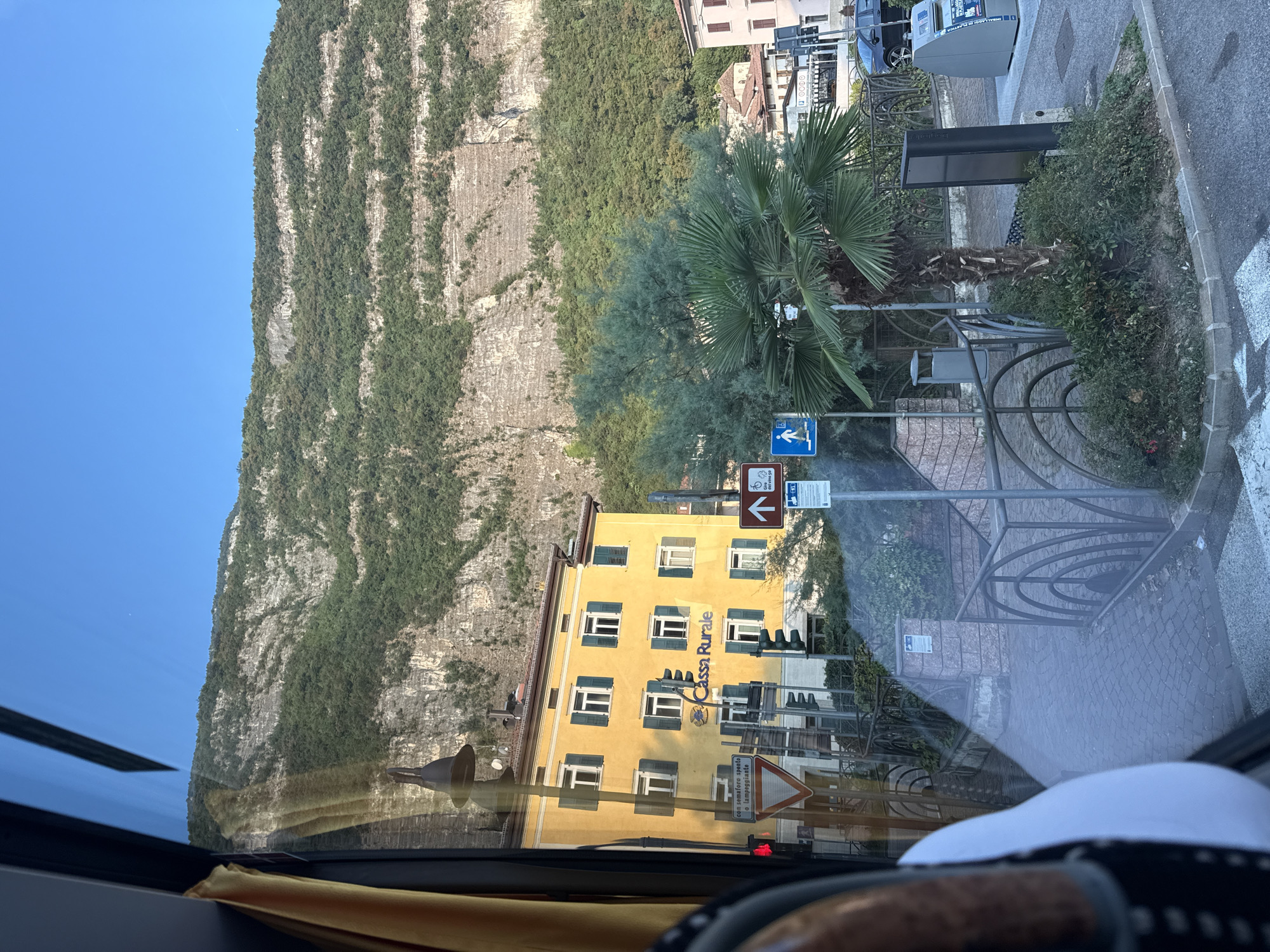 The image is a street view of Mezzolombardo, Italy, captured from inside a vehicle. A bright yellow building with green shutters is prominently featured, bearing the signage "Cassa Rurale". Adjacent to the building are traffic lights and various road signs, including one with a pedestrian symbol and another indicating a bicycle path. A palm tree and other greenery are visible in the foreground, with a steep, rocky mountain covered in trees serving as the backdrop. The sky is a clear blue. The scene appears to be during daylight hours.