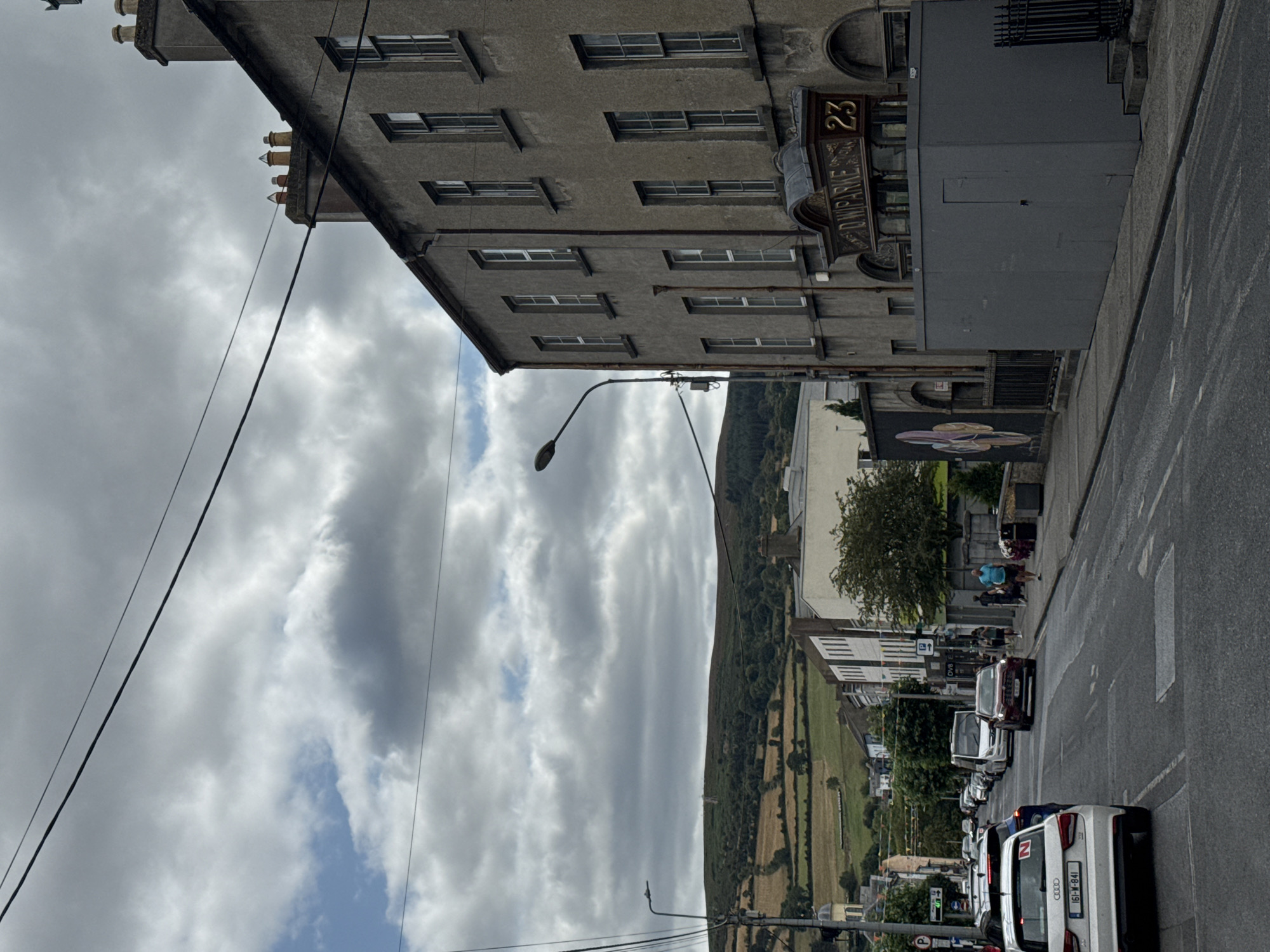 The image shows a street scene in Clonmel, Ireland, under a cloudy sky. The street is lined with buildings, some of which appear to be older stone structures, while others are more modern. There is traffic on the street, including cars, a van, and a bus. Pedestrians are visible on the sidewalks, and some are interacting with each other. In the background, there are rolling green hills covered in trees and fields. One building has signage that reads "D.W. PARKE" and "23". A mural is visible on the side of another building, depicting abstract colorful shapes. The overall impression is of a busy town on a somewhat overcast day.
