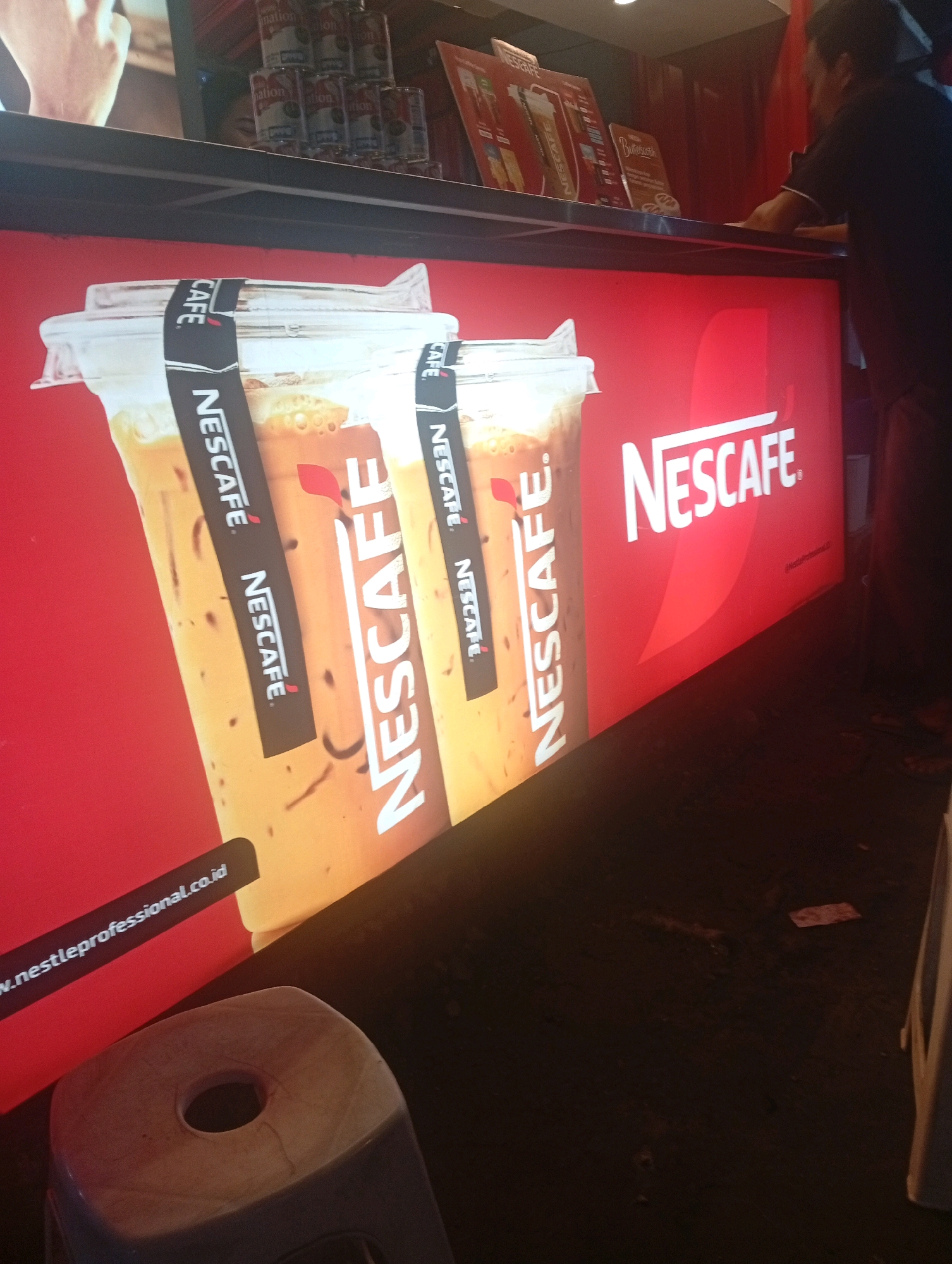 The image depicts a brightly lit counter display at a Nescafé cafe, likely in Banowan, Indonesia. The primary focus is on a large, illuminated advertisement featuring two tall Nescafé iced coffee drinks in clear plastic cups. The advertisement is predominantly red and white, with the Nescafé logo prominently displayed in white lettering. Black bands with the Nescafé logo and text are wrapped around the cups in the advertisement. Below the cups, a black banner with white text reads "www.nestleprofessional.co.id".
In the background, there are glimpses of stacked cans, possibly of Nescafé products, and menu boards displaying Nescafé offerings. A person, partially visible and out of focus, wearing a dark t-shirt, is standing behind the counter, interacting with the display. The lower portion of the image shows a dark floor and a corner of a white plastic stool. The overall impression is that of a commercial establishment advertising Nescafé beverages.