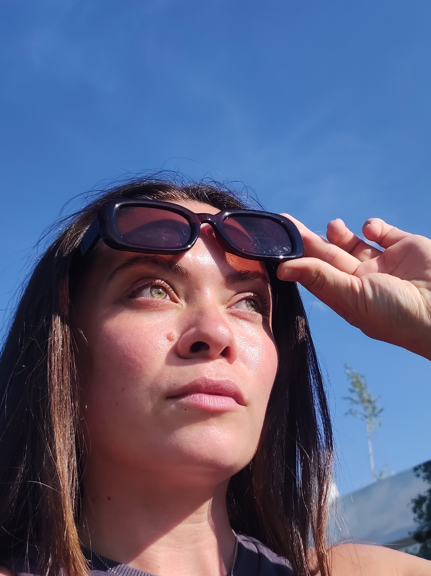 A young woman with long, dark hair is looking upwards, holding black sunglasses on her forehead. She has light skin with a dusting of freckles and her eyes are a striking green. The bright sun casts a shadow across the right side of her face, highlighting the contours of her cheek and nose. Her lips are slightly parted, and she appears to be squinting, as if shielding her eyes from the sun. She is wearing a dark-colored top, and a sliver of a silver chain is visible around her neck. In the background, the sky is a clear, vibrant blue, with the top of a slender, green plant visible on the right. A blurred, white structure with a darker shadow is also present in the far right background. The image was taken outdoors, likely on a sunny day. There is no indication of the city of Chaikovsky, Russia, in the image.