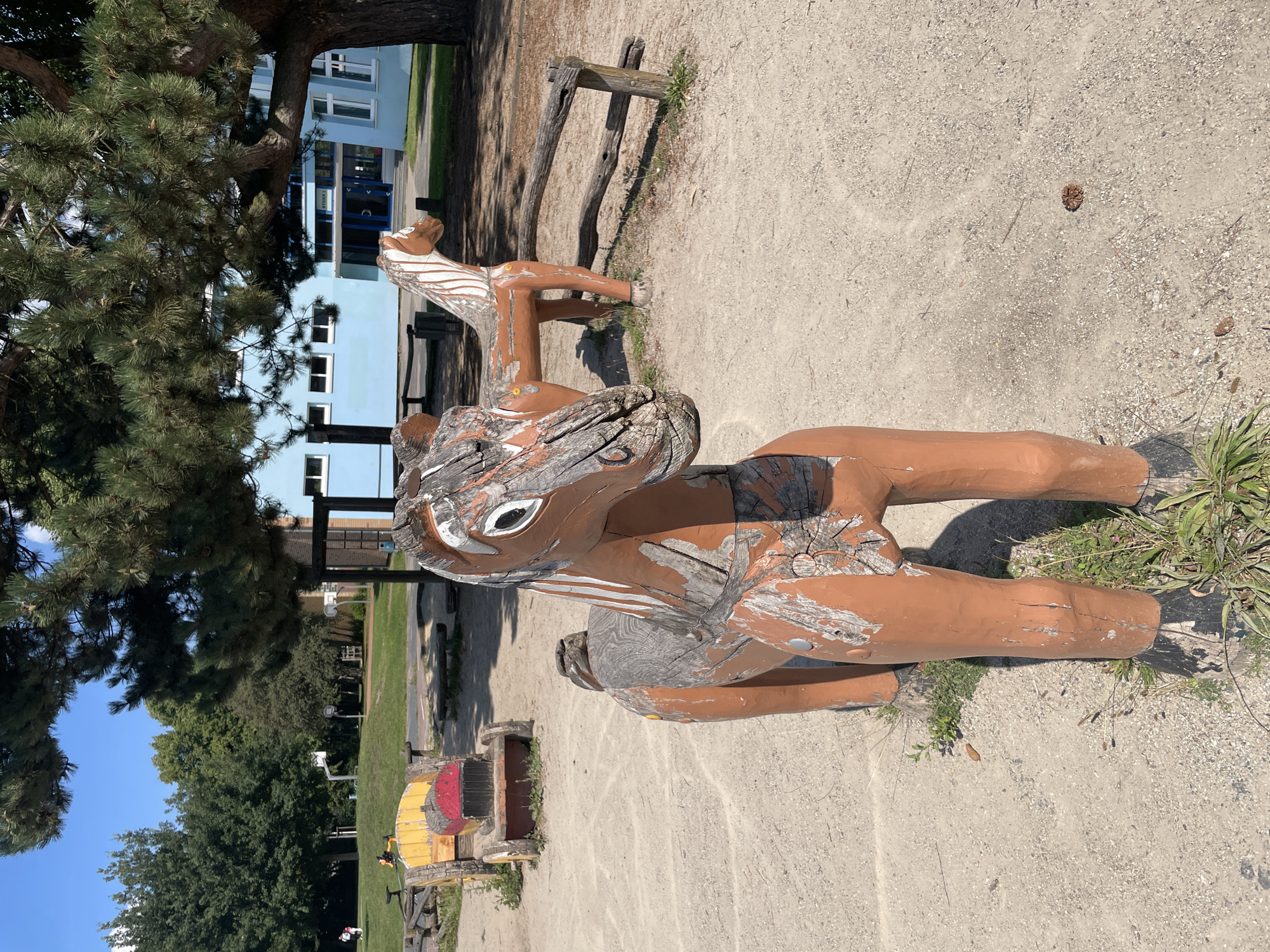 A sunny outdoor scene depicts a playground area with wooden horse sculptures as the foreground subjects. The closest horse sculpture is a weathered, brown wooden depiction of a horse's head and upper body, facing to the right. It has white painted eyes with black pupils and white painted stripes on its neck. The wood is cracked and peeling, revealing a lighter wood underneath. Behind this sculpture, another similar wooden horse sculpture is visible, also facing to the right, and appears to be a bench or ride-on toy.
In the background, to the left, a small yellow and red wooden cart-like structure with wheels is visible. Beyond the playground, a light blue building with multiple windows and an entrance is situated. The ground is covered with gravel and dirt, with patches of grass growing around the sculptures. Trees, including a large pine tree with dense green needles, are present in the background, casting shadows on the ground. The sky is clear and blue. The scene appears to be a public park or schoolyard. The image was taken in Bremerhaven, Germany.