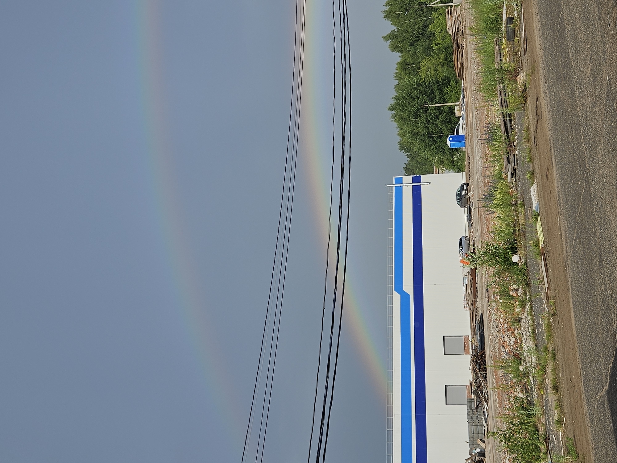 The image depicts a construction or demolition site in Chaikovsky, Russia, under a cloudy sky where a double rainbow is visible. In the foreground, there is a paved area and then a rough, unpaved ground covered with debris, including bricks, wood, and metal scraps. Vegetation, such as grass and some flowering plants, is growing throughout the debris-strewn area.
In the middle ground, a modern, white building with blue trim is situated. Two large, grey, shuttered windows are visible on its facade. In front of the building, there are piles of construction materials. Several vehicles are present: a silver car and a black car are parked near the building, and a portable toilet is to the right of the black car. Behind the building, a dense line of green trees forms the background. Overhead, black power lines stretch across the frame.
The double rainbow arcs across the sky, with the primary rainbow appearing more distinct than the secondary one above it. The overall impression is of a work in progress, possibly post-demolition or a construction site undergoing development, set against a backdrop of a brief weather event.