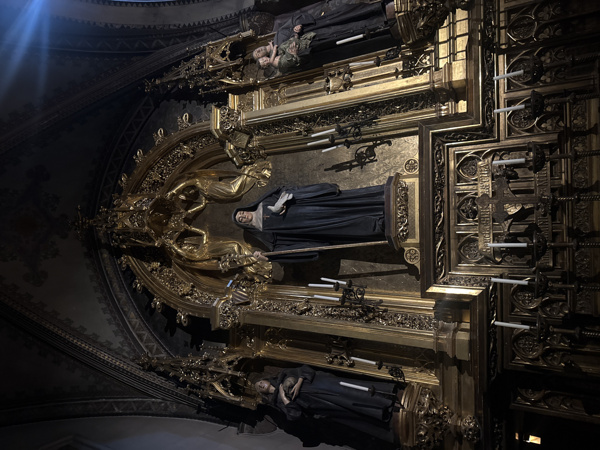 The image shows an ornate altar in a church, likely within the city of Monistrol de Montserrat, Spain, given its proximity to the Montserrat Monastery. The altar is predominantly gold and features elaborate carvings and statues.
In the center of the altar, a statue of a nun in dark robes stands holding a staff and a book. To her left, another statue depicts a nun holding a skull. To the right of the central nun, a statue shows a man holding a child. The statues are positioned on pedestals adorned with intricate golden detailing.
Several white candles are placed on candlestick holders around the altar, some lit and some unlit, casting a dim glow on the scene. The background of the altar is a rich gold, decorated with a patterned tapestry or relief. Above the central figures, golden architectural elements and angelic figures are visible, contributing to the grandeur of the setting. The ceiling of the church arches overhead, with dark, patterned decoration. The overall impression is one of religious solemnity and artistic opulence.