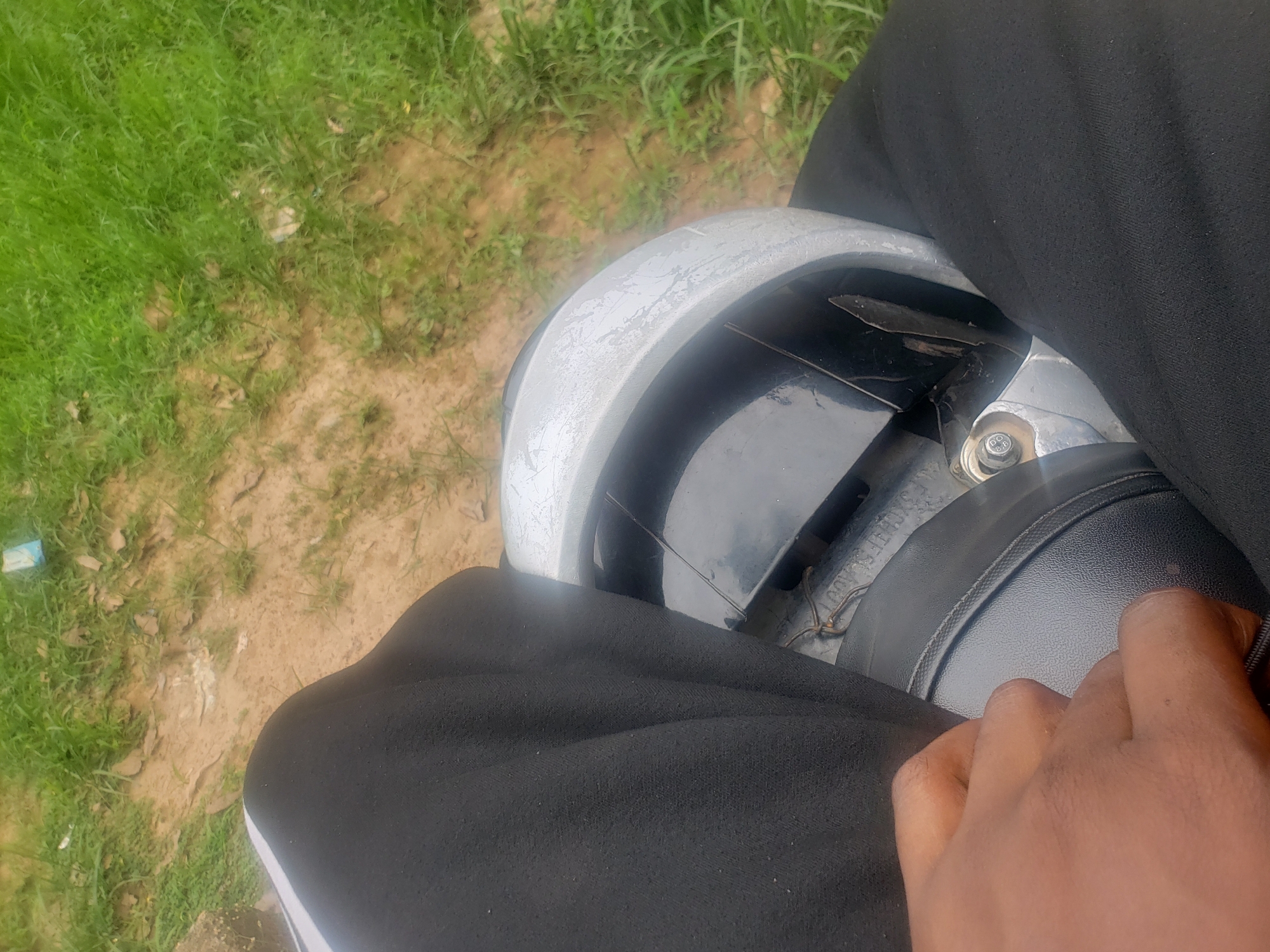 In a ground-level, first-person perspective shot taken in the city of Kaduna, Nigeria, a person's dark-skinned hand is visible, resting on the black seat of a motorcycle. The person is wearing black track pants with a white stripe down the side. The motorcycle's silver-colored handlebar fairing, showing signs of wear and tear, is also in view. The background consists of a mix of dry, sandy earth and green grass, indicating an outdoor, possibly unpaved, area. A small blue object, possibly a discarded wrapper, is visible in the grassy area. The lighting suggests it is daytime, with no harsh shadows indicating direct overhead sun.