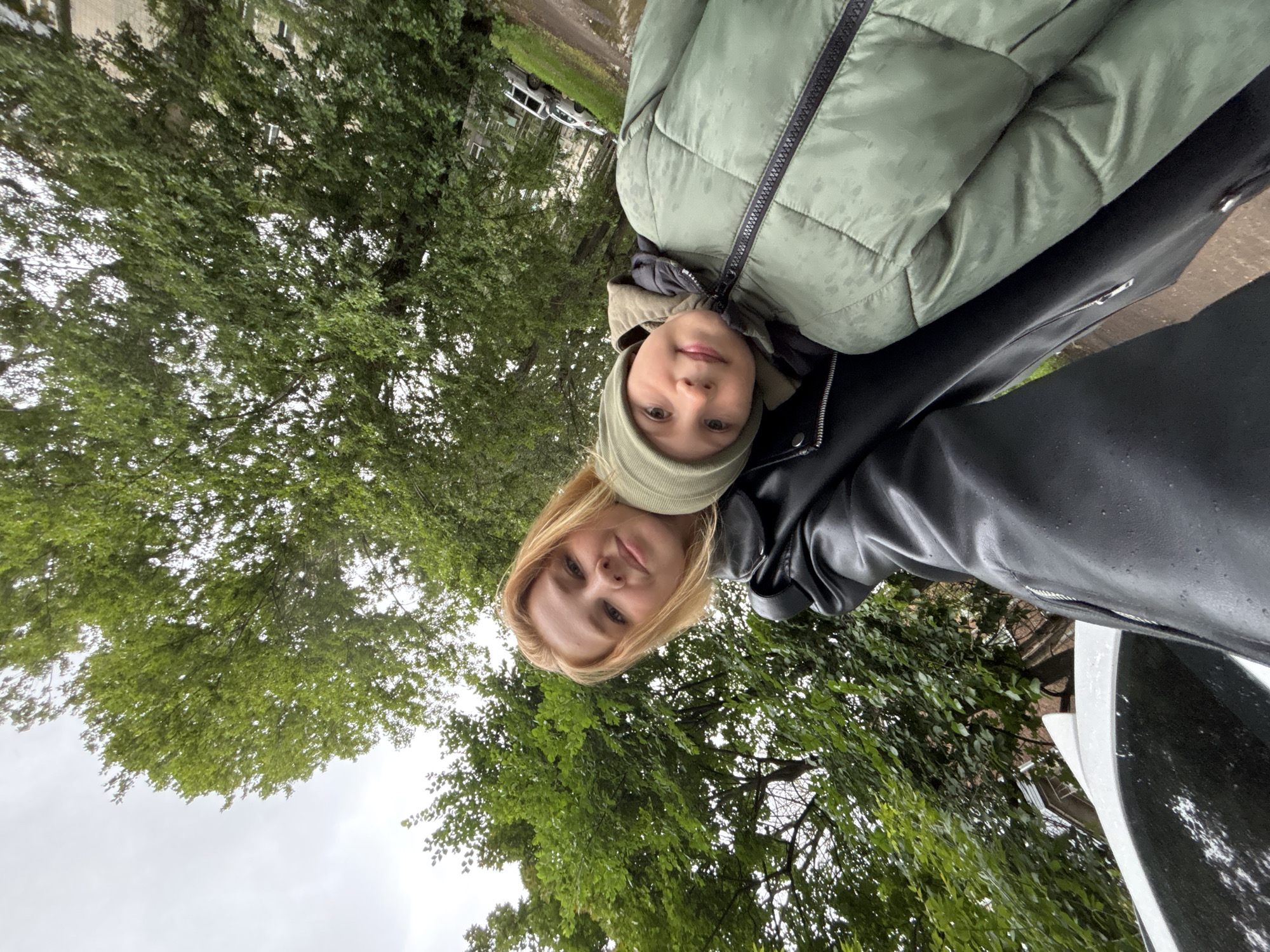 A woman and a child are taking a selfie outdoors, with lush green trees framing the shot. The woman, with short blonde hair, wears a black leather jacket. The child, wearing a green hooded jacket and a matching hat, is nestled close to her. In the background, a white car is parked on a wet, paved area, suggesting it may have recently rained. The surroundings appear to be a residential area with buildings visible further in the distance. The overall atmosphere is casual and intimate, capturing a moment between the two individuals. The location is identified as Rusanovo, Russia.