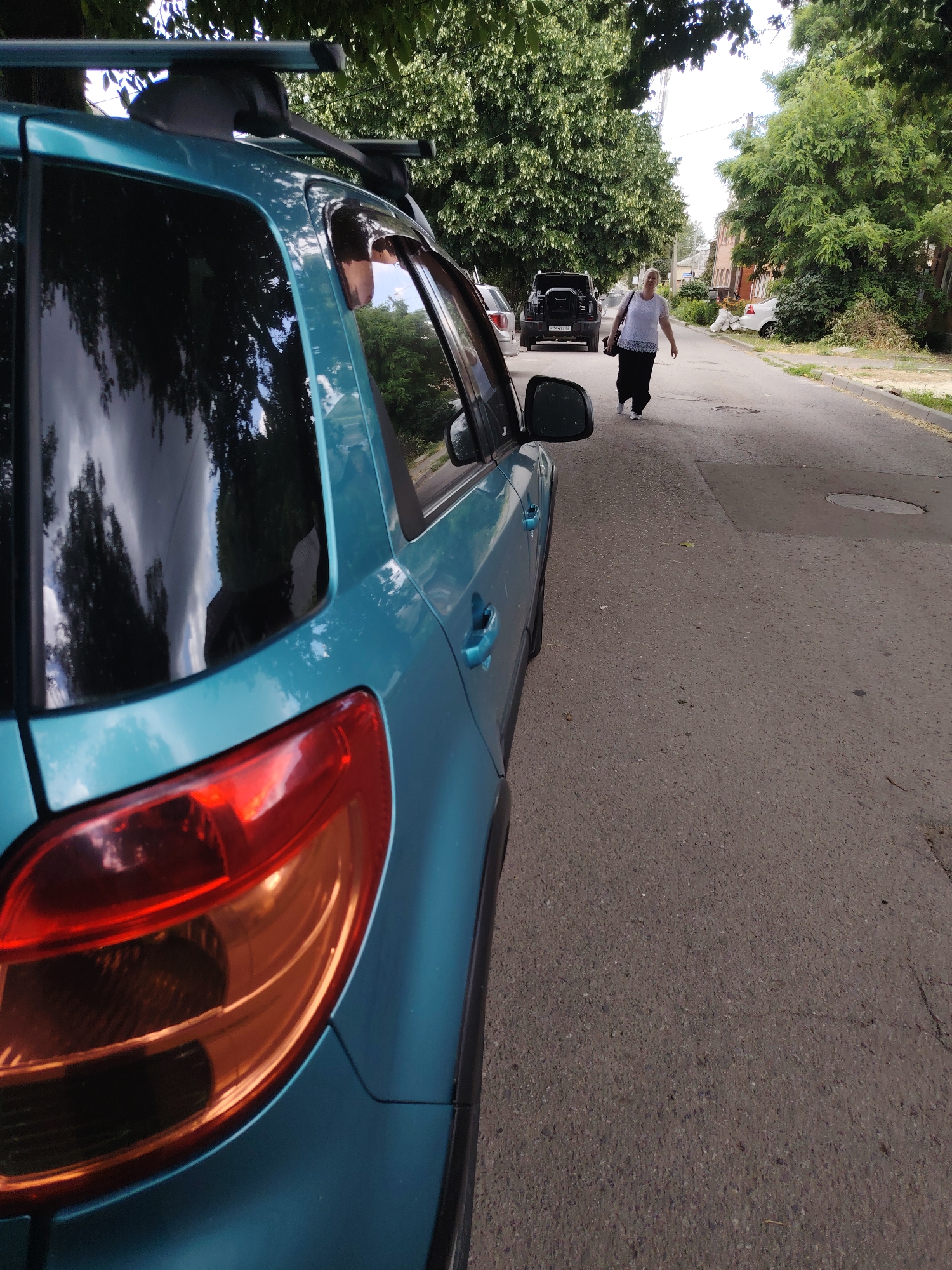 A woman, wearing a white t-shirt and black pants, walks down a paved street lined with trees. She is carrying a dark bag over her shoulder. A teal-colored car is parked on the left side of the street, with its rear visible in the foreground. In the background, several other vehicles are parked, including a black SUV. The scene appears to be in a residential area, with buildings visible on the right side of the street. The weather seems to be overcast, and the time of day is likely daytime. The overall mood is casual and ordinary. Some visible text on the black SUV's license plate reads "M473YX 762."