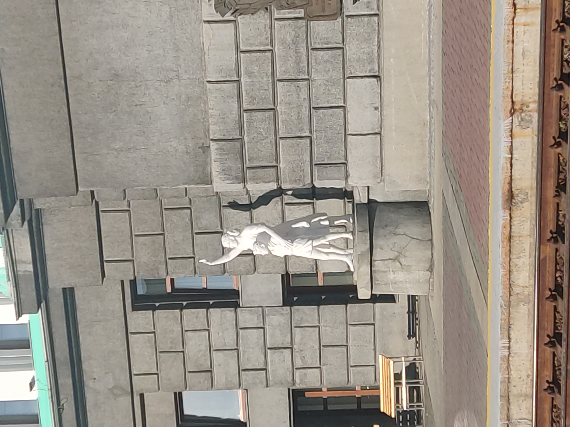 The image shows a close-up view of a building facade with a relief sculpture. The sculpture features two figures, appearing to be male and female, intertwined. The figures are white and appear to be made of stone or a similar material. The male figure is behind and above the female figure, with his arms around her. The female figure is reaching out with her left arm. The sculpture is mounted on a concrete pillar that is integrated into the building's architecture. The building itself is constructed with large, grey, rectangular blocks that resemble stone or concrete. There are windows visible on the building, some of which have dark frames. The overall aesthetic of the building and sculpture suggests a modernist architectural style. The scene is lit by daylight, casting a distinct shadow from the sculpture onto the building's surface, indicating it is likely daytime and sunny. The context provided states the location is Novosibirsk, Russia, which might offer further clues to the specific architectural style or historical significance of the building and sculpture, though no text is visible to confirm this. The activity depicted is a static representation in sculpture, not an ongoing event. The notable details include the artistic style of the relief, the materials used in construction, and the interplay of light and shadow on the facade.
