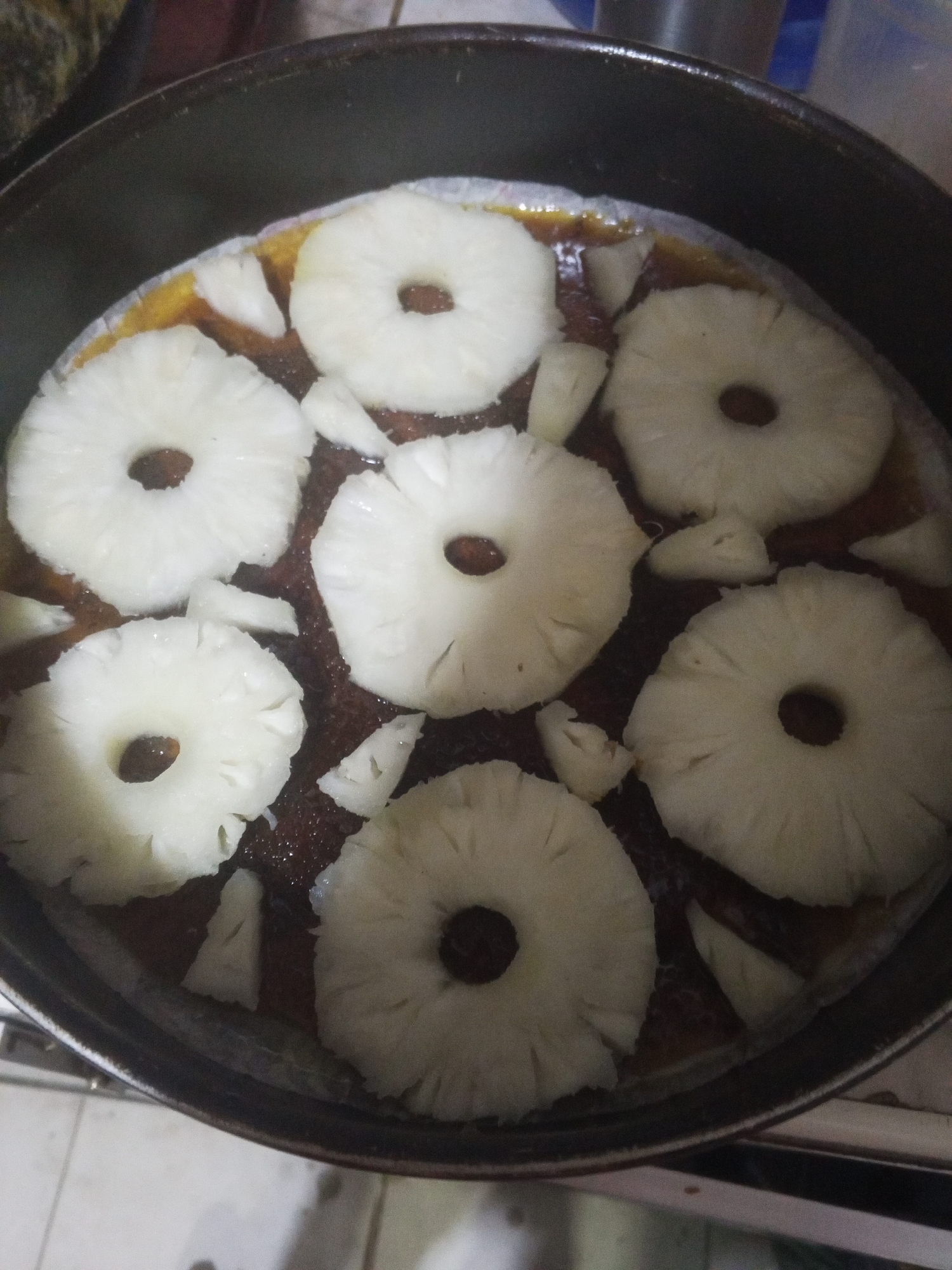 The image shows a pineapple upside-down cake in a dark-colored pan. The cake appears to be arranged with several pineapple rings, interspersed with smaller chunks of pineapple, sitting on top of a darker, possibly caramelized base. The photo was taken indoors in Tinaquillo, Venezuela.