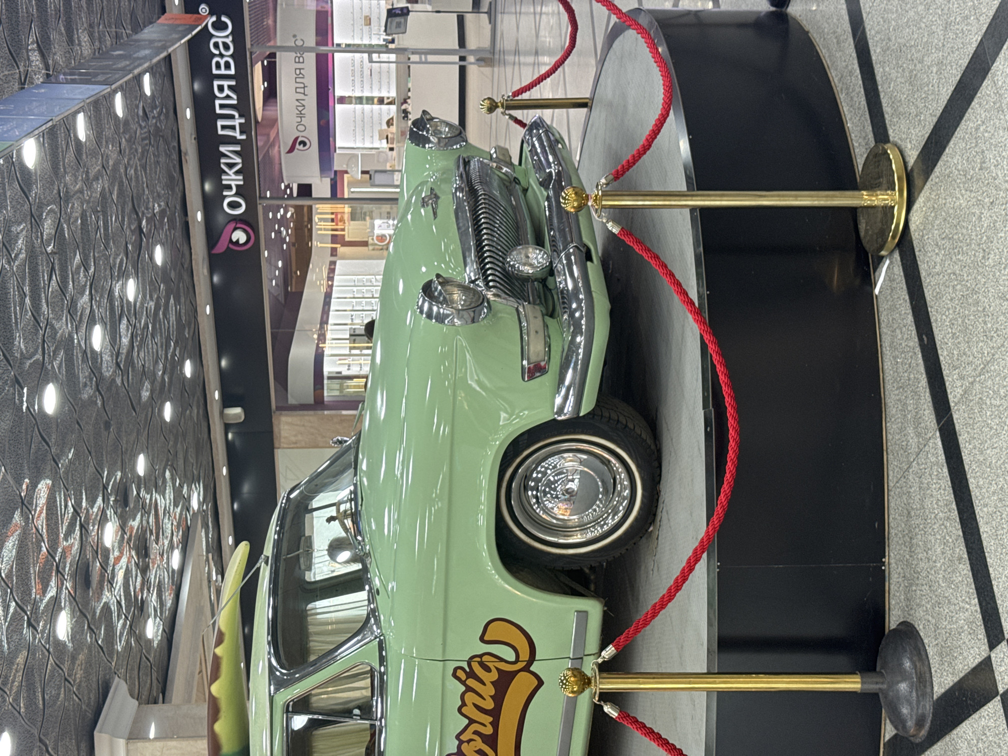 A vintage, light green GAZ-21 Volga car is displayed on a circular black platform, roped off with red cord and gold stanchions. The car features a "California" decal on its side and what appears to be a surfboard secured to its roof. The background reveals a modern shopping mall interior with a patterned ceiling, recessed lighting, and storefronts. Notably, signage for "очки для вас" (meaning "glasses for you") is visible, indicating an optician's store. The flooring is a patterned tile. No individuals are clearly depicted in the immediate foreground or interacting with the car. The scene is indoors, consistent with a public exhibition or display within a commercial space in Yekaterinburg, Russia.
