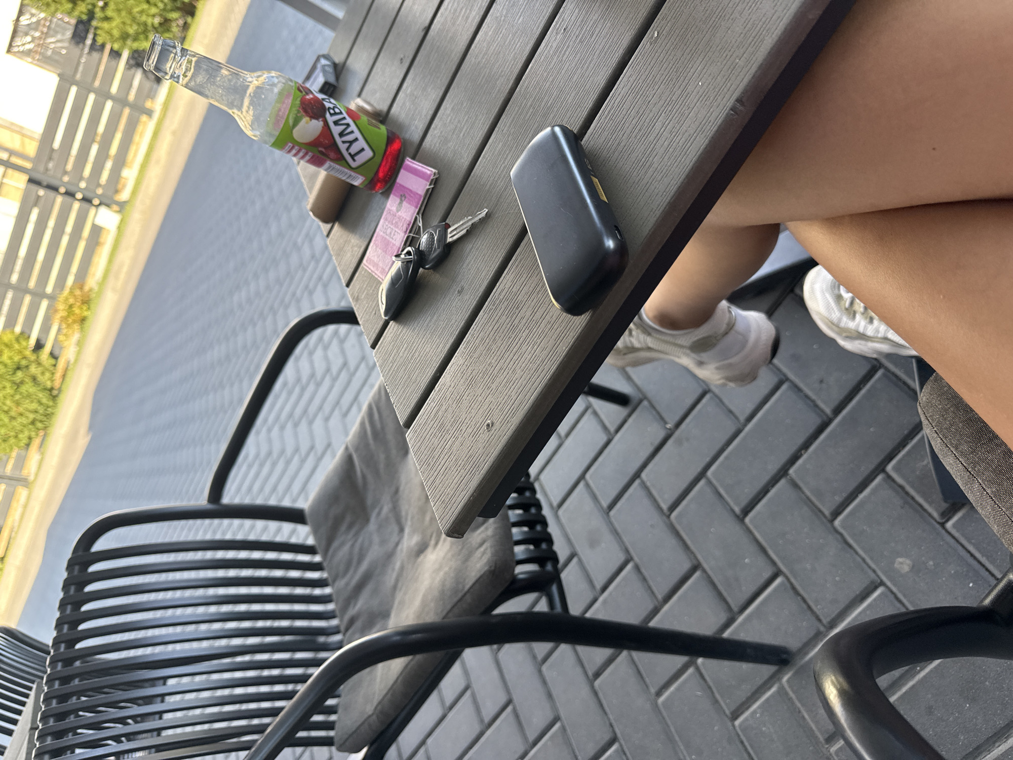 A person is sitting at an outdoor table in Comrat, Moldova. The table is made of dark wood slats and has a few objects on it: a glass bottle with a red liquid and a green label that says "TYMBA", a set of car keys, a pink object that appears to be a napkin or receipt, and a black electronic device. The person's legs are visible, wearing white sneakers and a dark gray skirt. In the background, there is a black chair with a gray cushion and a paved ground made of dark gray rectangular tiles. A light-colored fence and green bushes are visible in the far background. The scene appears to be in daylight, possibly late afternoon, given the lighting.