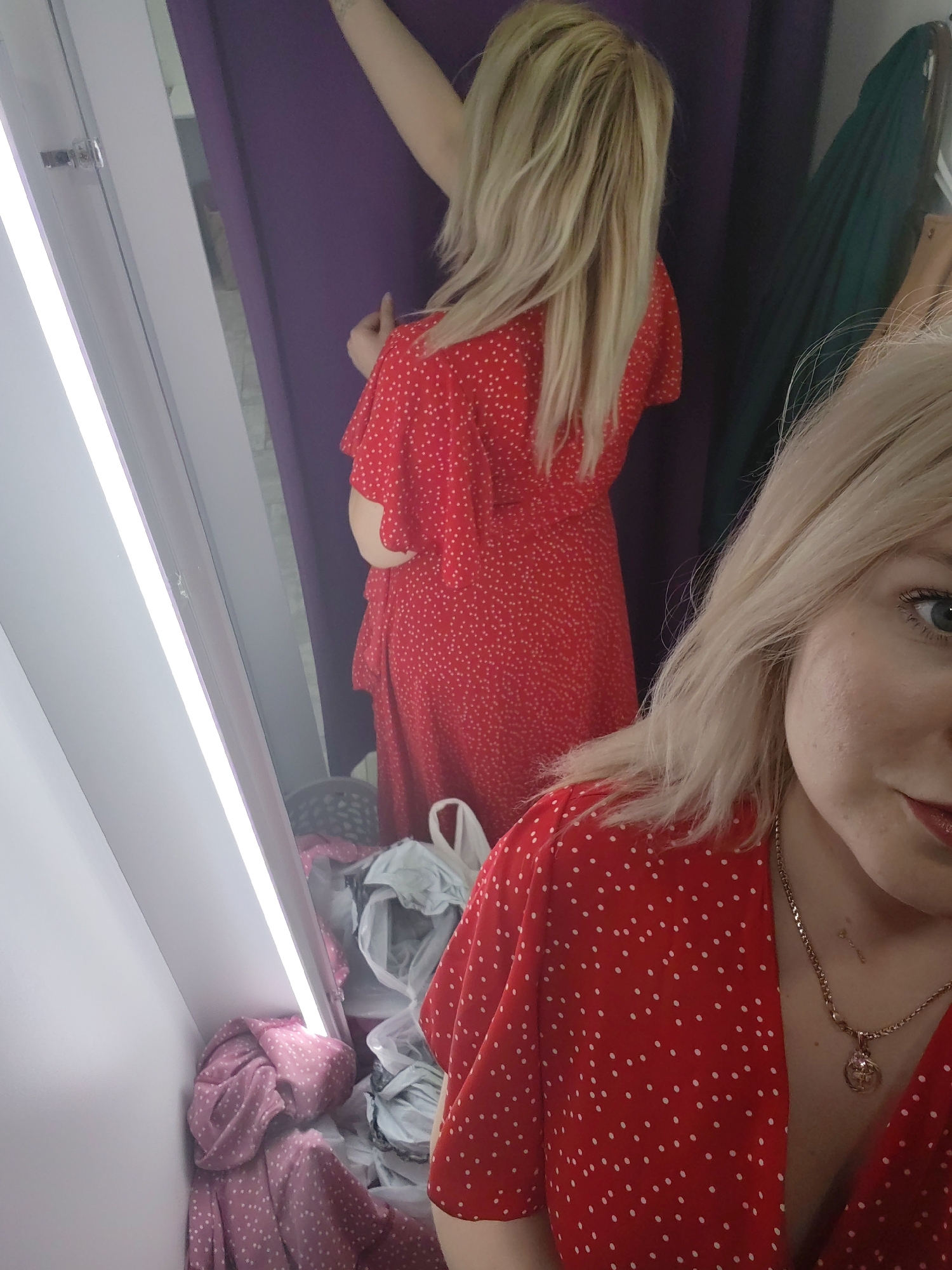 A blonde woman takes a mirror selfie in what appears to be a changing room in Izhevsk, Russia. In the foreground, the right side of her face is visible, with fair skin, a light blue eye, and red-painted lips hinting at a slight smile. Her long, blonde hair cascades over her shoulder. She wears a red dress with small white polka dots, and a silver chain necklace with a circular pendant rests on her décolletage.
The main subject is her reflection in the mirror, showing her full figure. She is wearing the same red polka-dot dress, which has short, loose sleeves and a cinched waist. Her long blonde hair is visible from behind. Her right arm is raised, holding back a purple changing room curtain, which frames the background behind her reflection. To the right of her reflection, partially obscured by the curtain, a dark green garment hangs.
The changing room environment is evident from the large mirror, the purple privacy curtain, and a bright, vertical light strip on the left side of the mirror, providing illumination. At the bottom of the mirror's reflection, a pile of discarded or untried clothes, including a light pink polka-dot item, and shopping bags are visible on the floor. The scene captures the woman trying on clothes and posing to view the outfit.