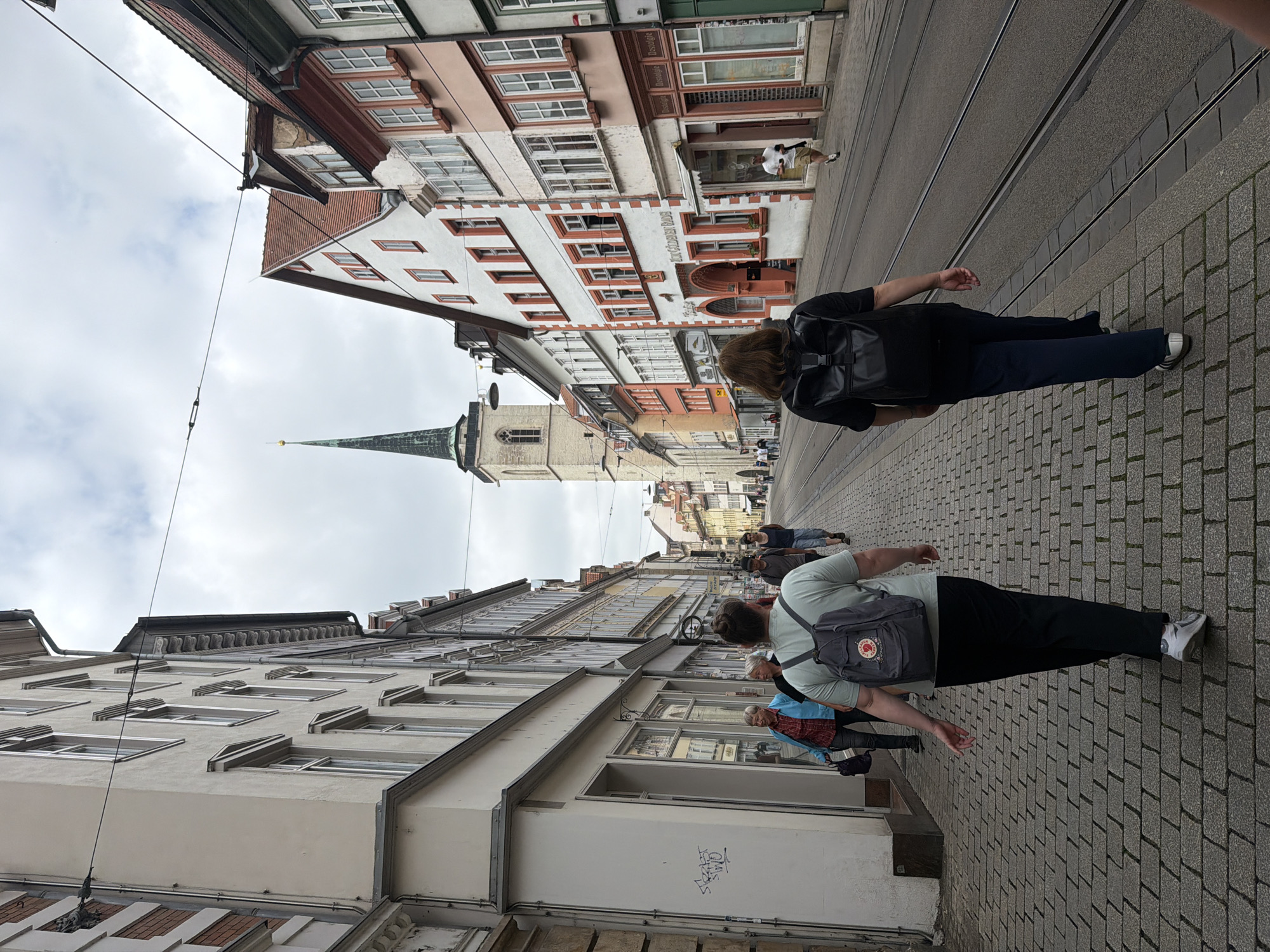 The image shows a street scene in Erfurt, Germany. Two women are walking away from the camera on a cobblestone sidewalk. The woman on the left has a light green t-shirt and a gray backpack, while the woman on the right has a black t-shirt and a black backpack. They are walking on a sidewalk next to tram tracks.
In the background, there are buildings with a mix of architectural styles. The most prominent structure is a tall church tower with a green spire, characteristic of churches in Erfurt. The buildings lining the street are colorful, with red, orange, and white facades. Some of the buildings appear to be shops with large windows displaying goods.
Other people are visible in the distance, walking on the sidewalk and in the street. The sky is overcast, suggesting a cloudy day. The overall scene depicts a typical European city street with pedestrian activity and historical architecture.